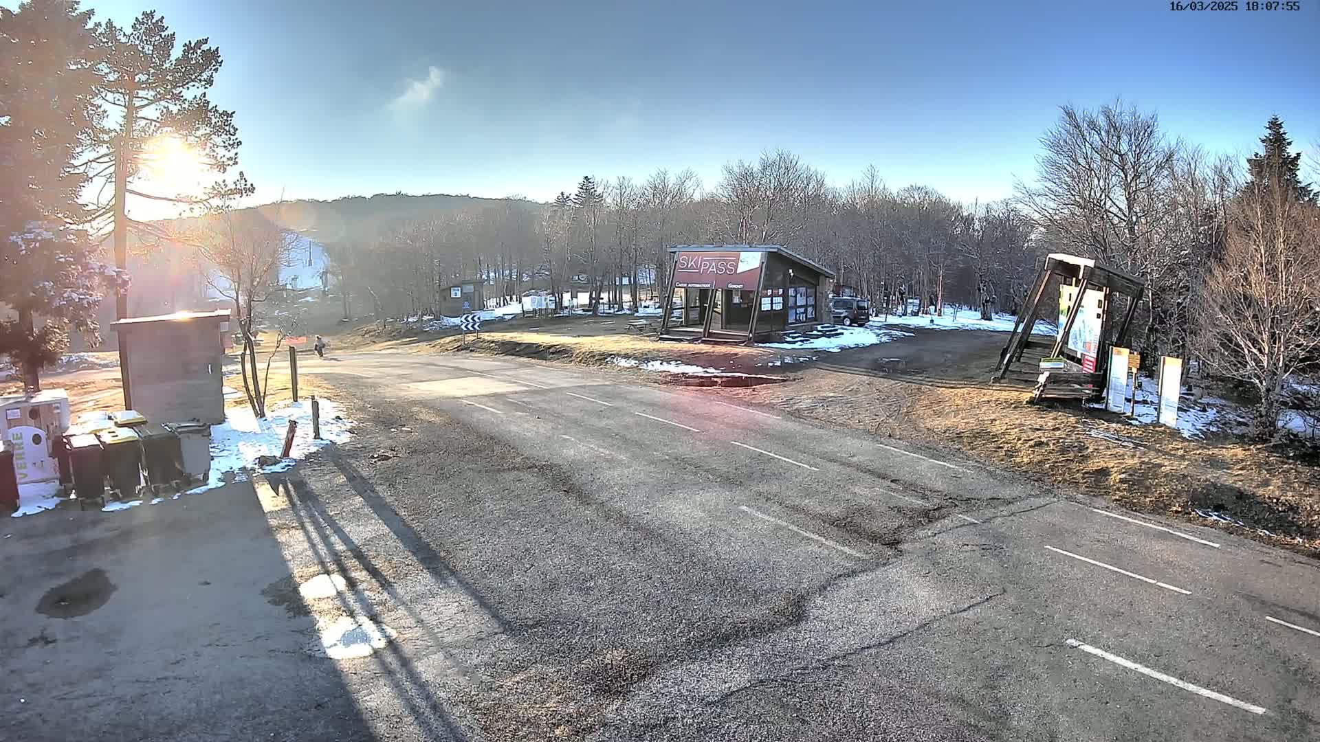 A sunny winter scene shows a paved road leading to a ski pass building and a ski slope in the background, with some patches of snow and bare trees.