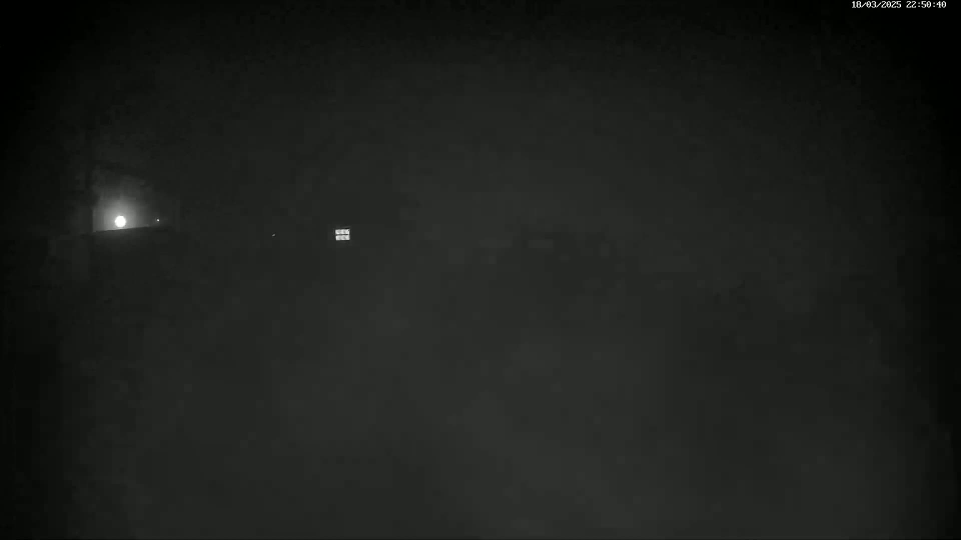 A dimly lit, nighttime scene shows a building with a lit window and a bright light possibly from a moon or lamp in the distance, under hazy or foggy conditions.