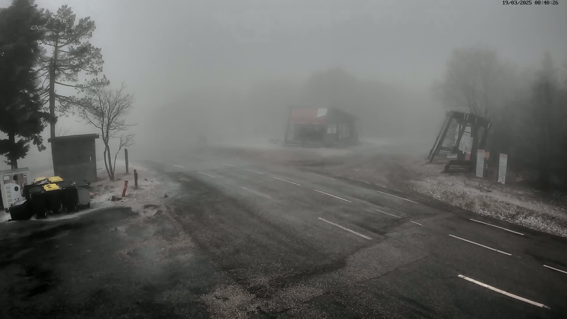A road intersection with a few buildings is shrouded in thick fog, with some snow or ice visible on the ground.