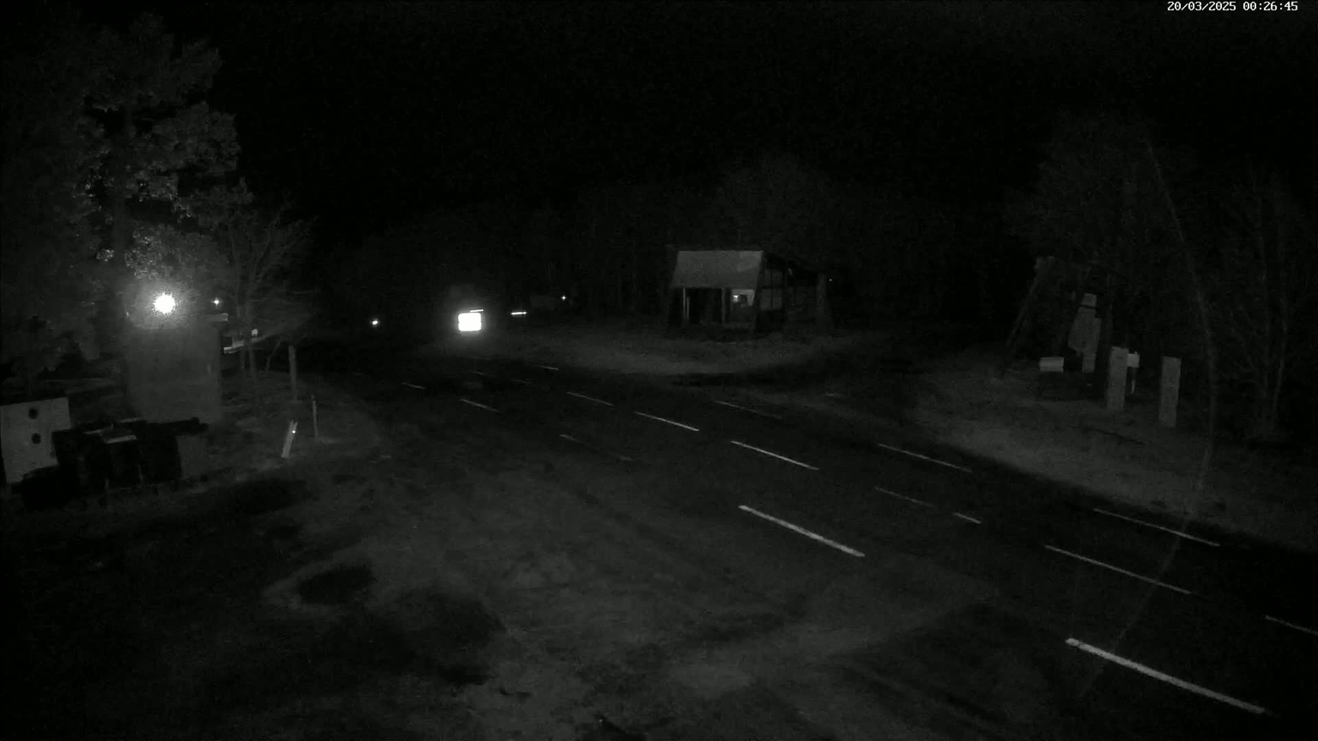 A dark, nighttime scene shows a sparsely trafficked road intersecting with another road, passing near a small building and some trees under a clear sky.