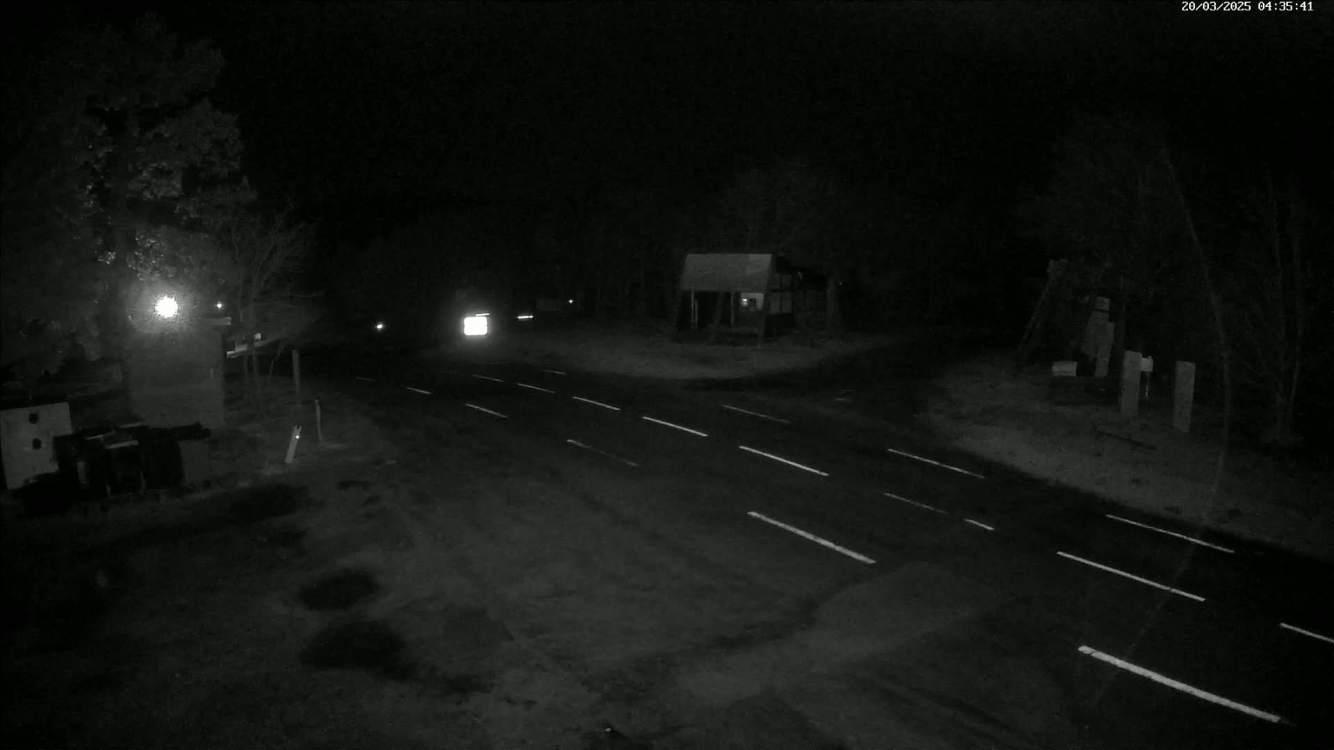 A dark, nighttime scene shows a sparsely trafficked road intersection with some trees and buildings visible in the distance.