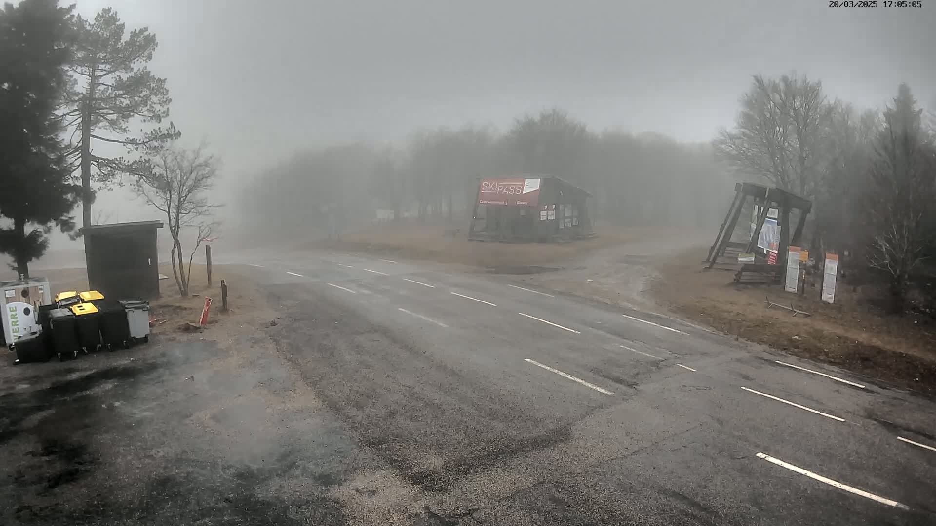A foggy, outdoor scene shows a paved road intersecting with another road near a small building and several trash receptacles, surrounded by leafless trees.