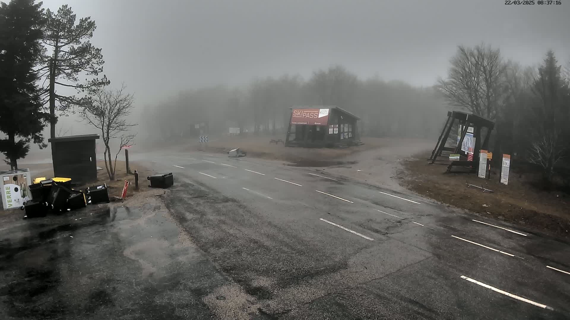 A wet road intersects in front of a small building in a foggy, outdoor setting, with several overturned trash cans scattered nearby.