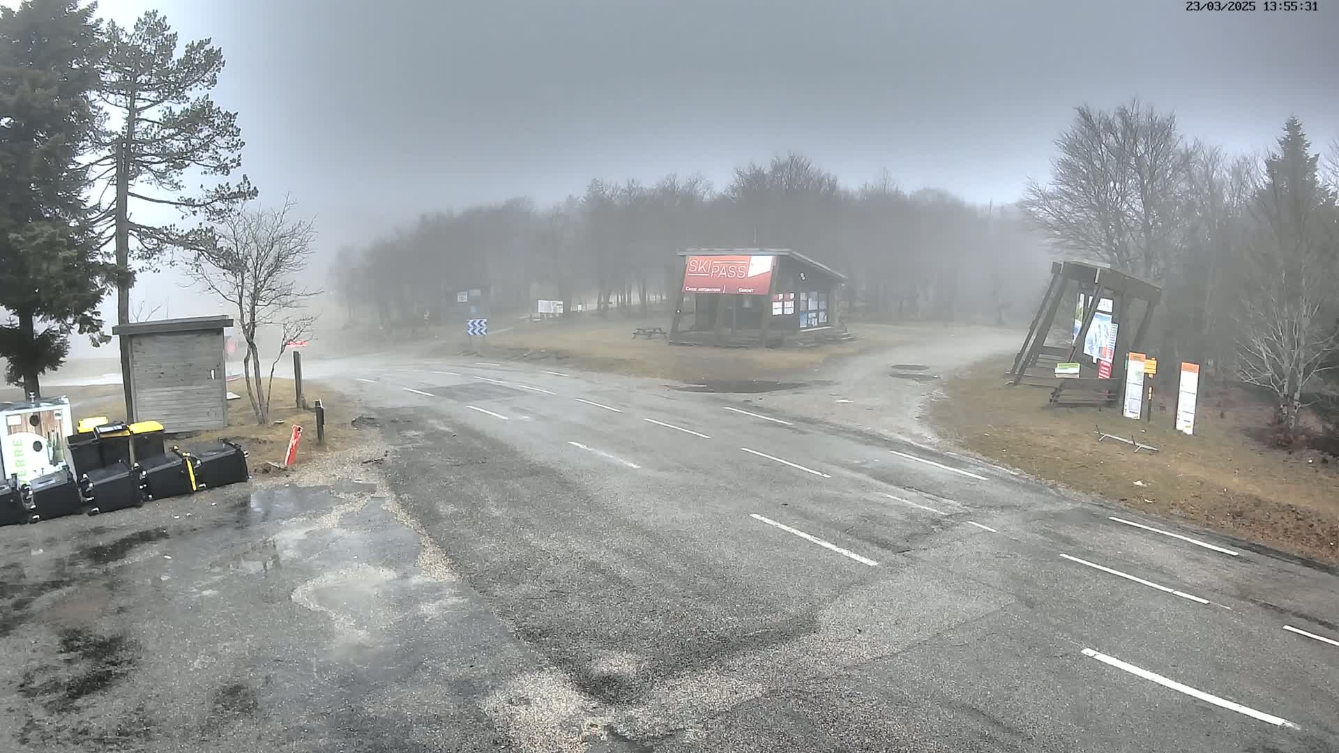 A foggy road intersection with a ski pass booth and several trash cans sits near a wooded area.