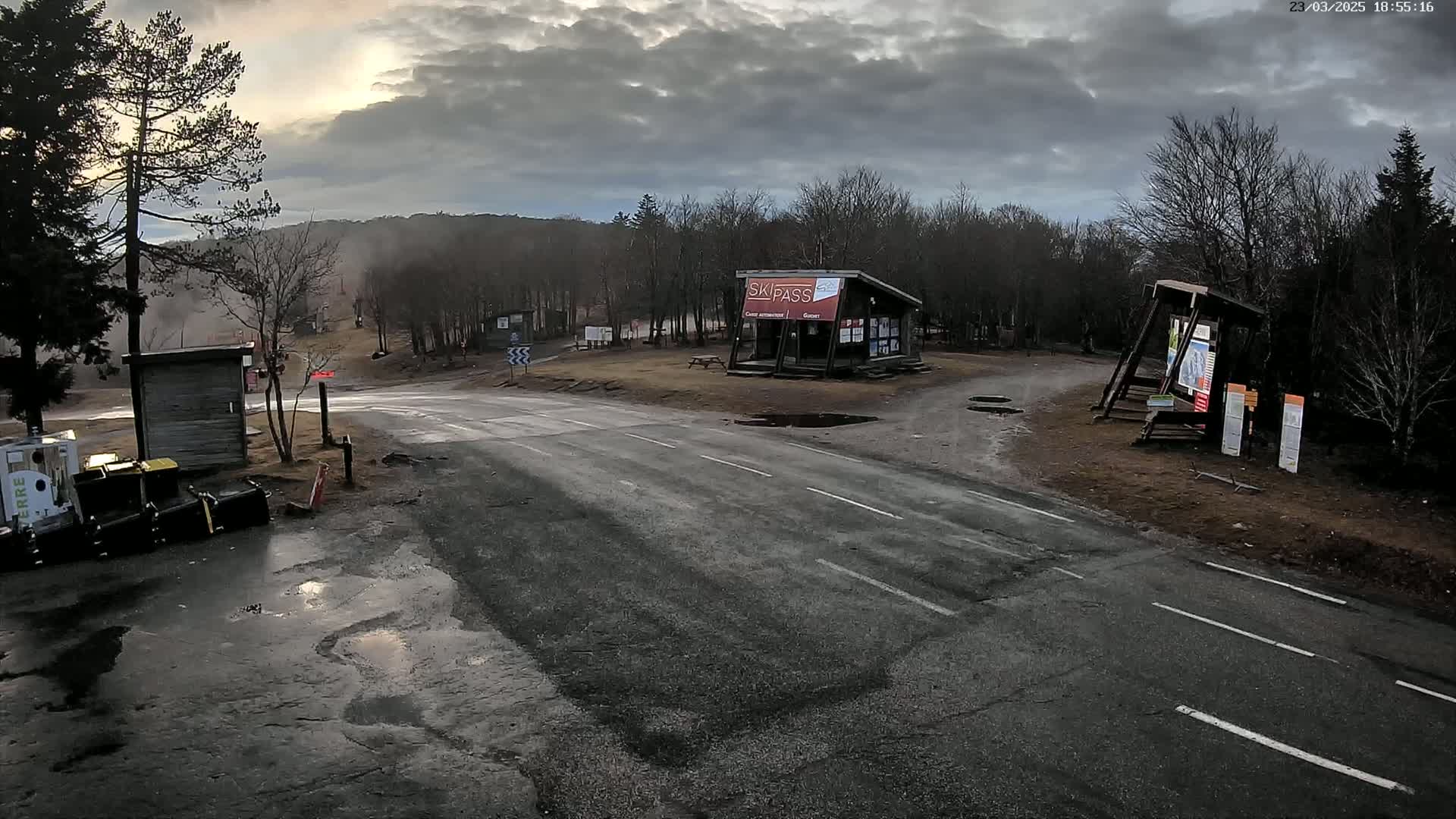 A wet asphalt road leads to a ski pass building and information kiosks in a wooded area under a cloudy sky.