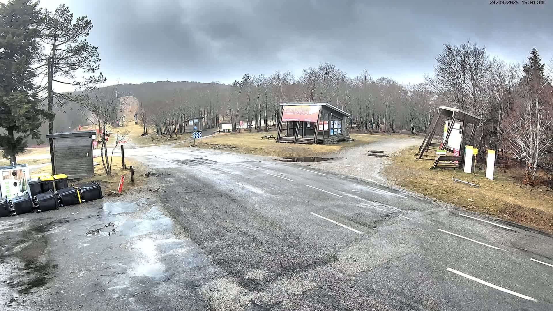 A wet asphalt road leads to a ski lift, flanked by small buildings and bare trees under an overcast sky.