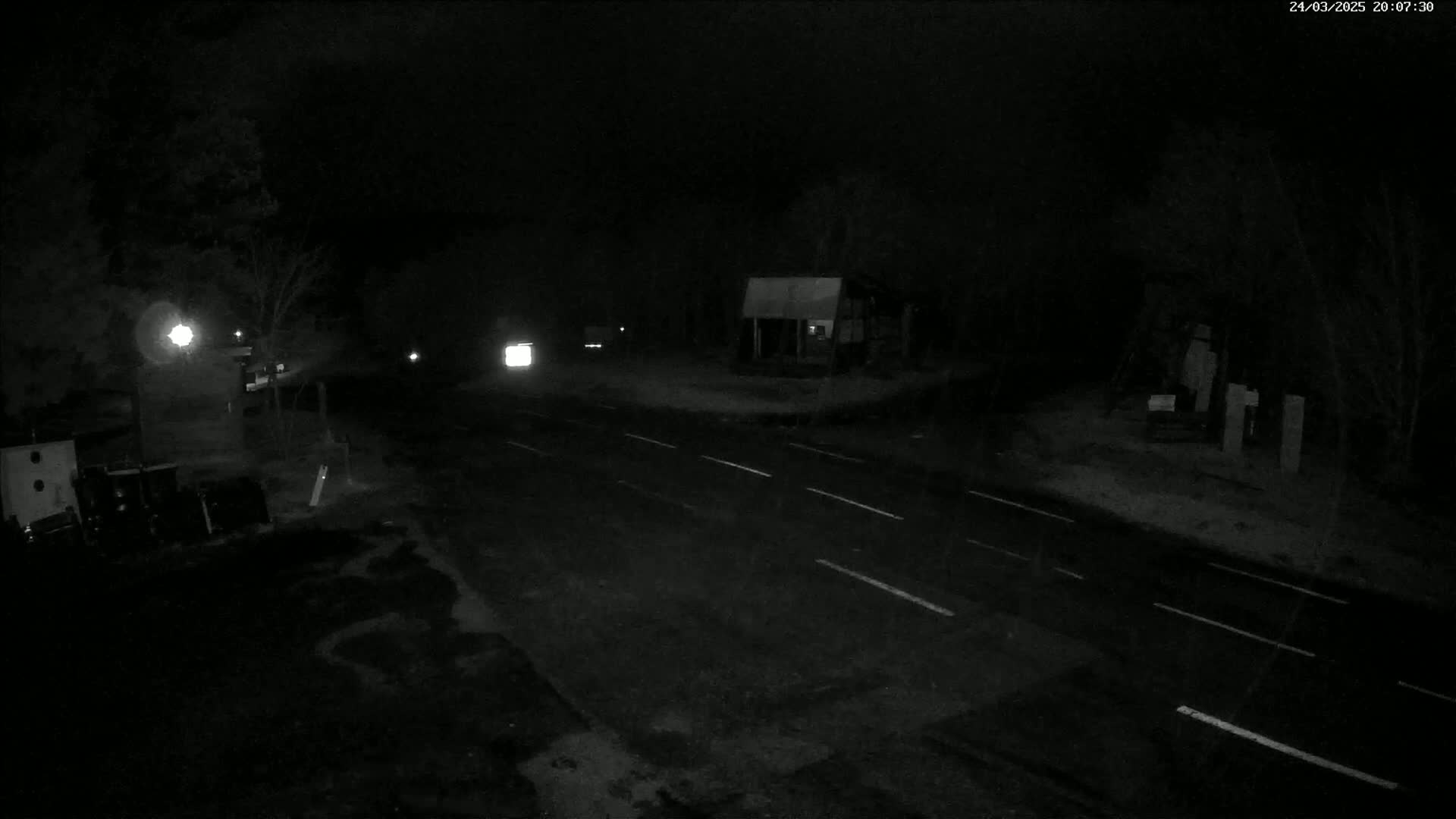 A dark, nighttime scene shows a sparsely traveled road with faint vehicle headlights in the distance, under snowy or rainy conditions.