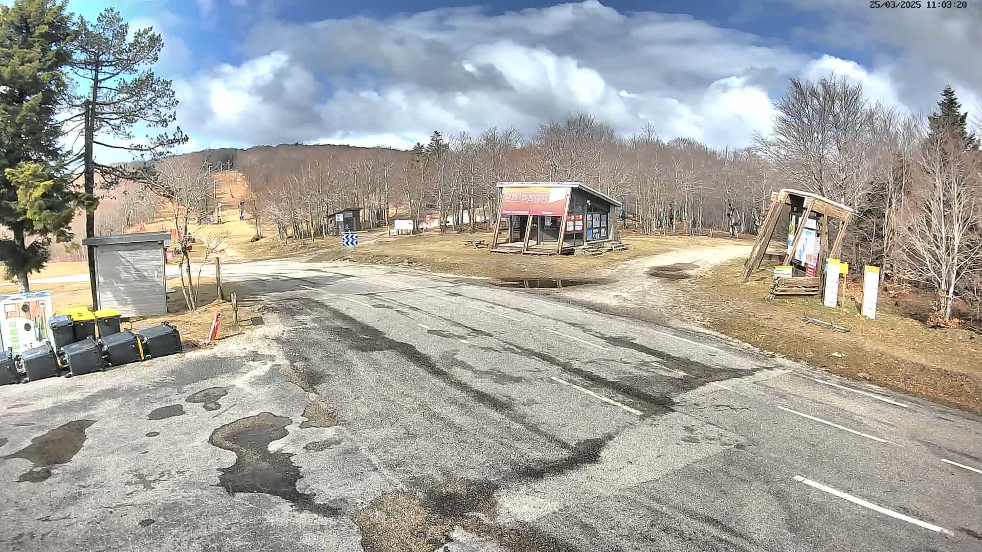 A mostly sunny day shows a paved road leading to a ski area with several small buildings and a sparsely populated, leafless forest in the background.