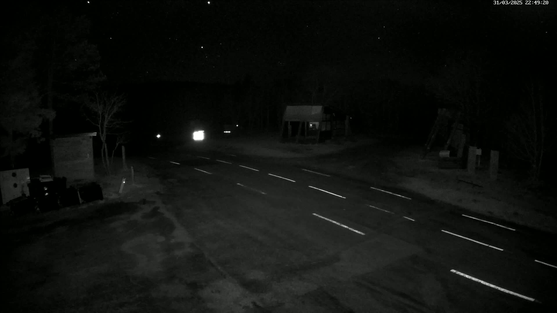 A nighttime, low-light image shows a dark road with white lane markers, a few vehicles with headlights on in the distance, and some sparse trees and small structures along the roadside under a clear, starry sky.