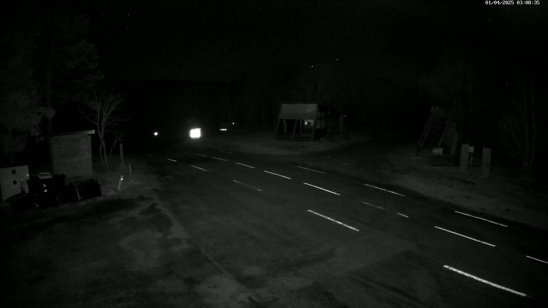 A dark, nighttime road scene shows a two-lane road with a vehicle's headlights visible in the distance,  surrounded by sparse trees and a small building under a clear, starry sky.