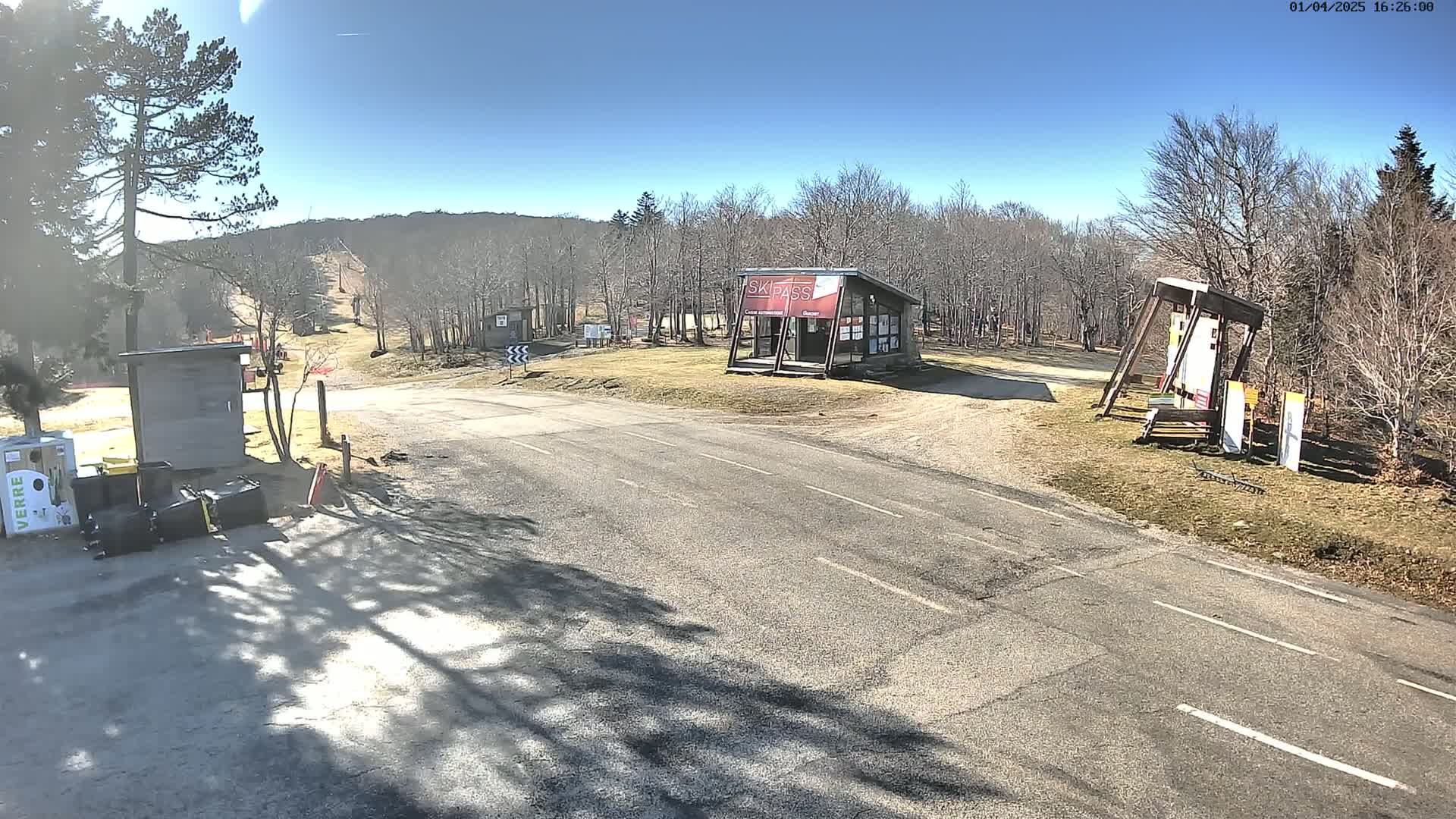 A paved road leads to a ski area entrance on a sunny day, with small buildings and signage visible, surrounded by bare deciduous trees and some evergreens.