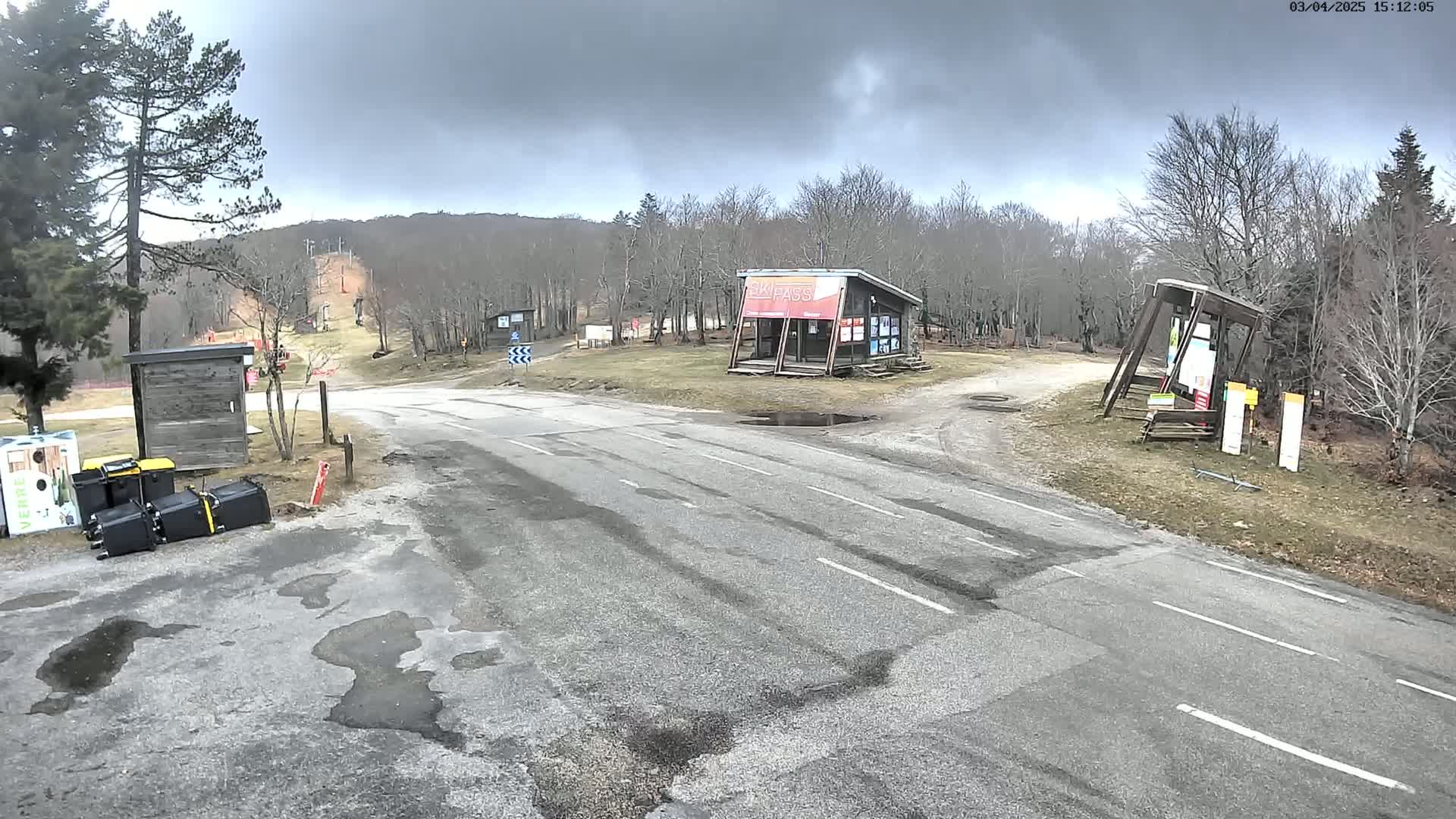 A paved road leads to a ski area under a cloudy sky, with several small buildings and a few scattered trash cans visible near the road.