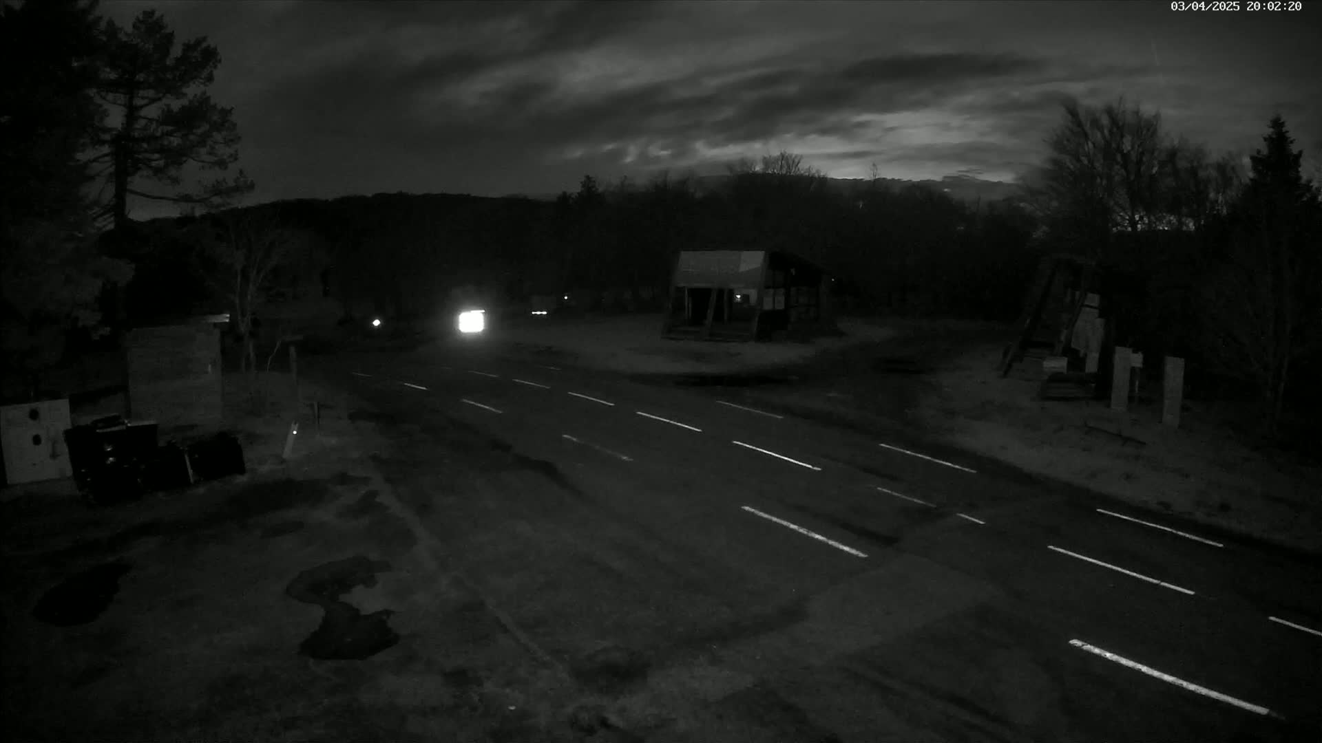 A nighttime, overcast scene shows a dark road with white lane markings, a vehicle's headlights in the distance, and a small building near some trees.