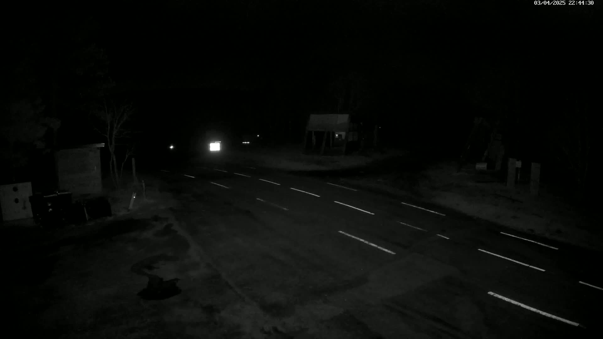 A dark, nighttime view of a sparsely lit road with a vehicle's headlights visible in the distance.