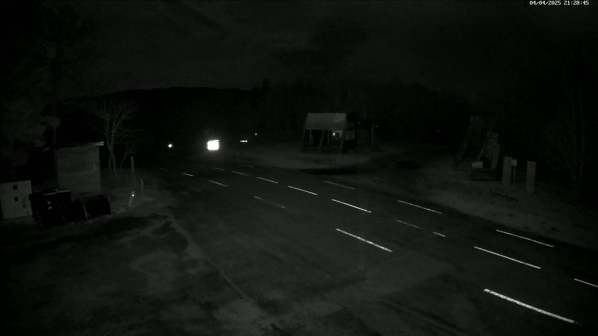 A dark, nighttime scene shows a sparsely lit road with a few vehicles and buildings in the distance, possibly with some snow or ice on the ground.