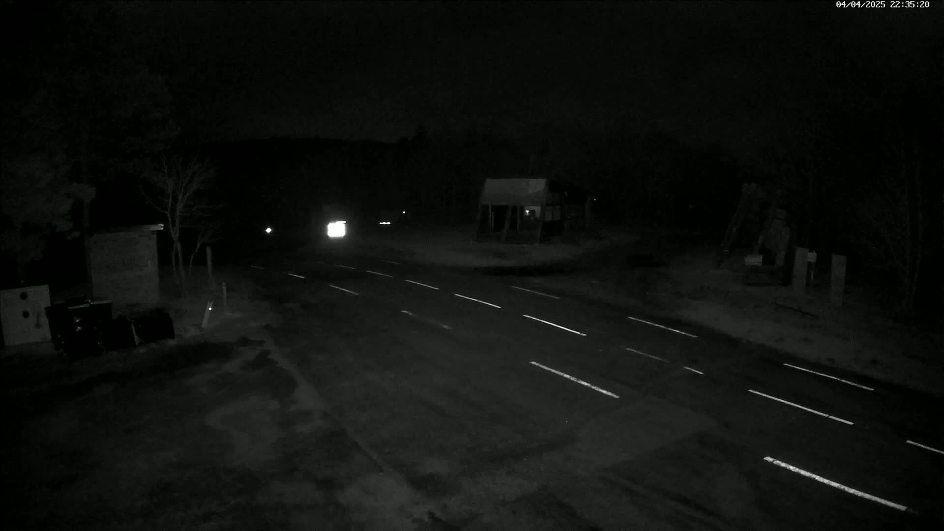 A nighttime, snow-covered road intersection with a few vehicles and small buildings visible under low-light conditions.