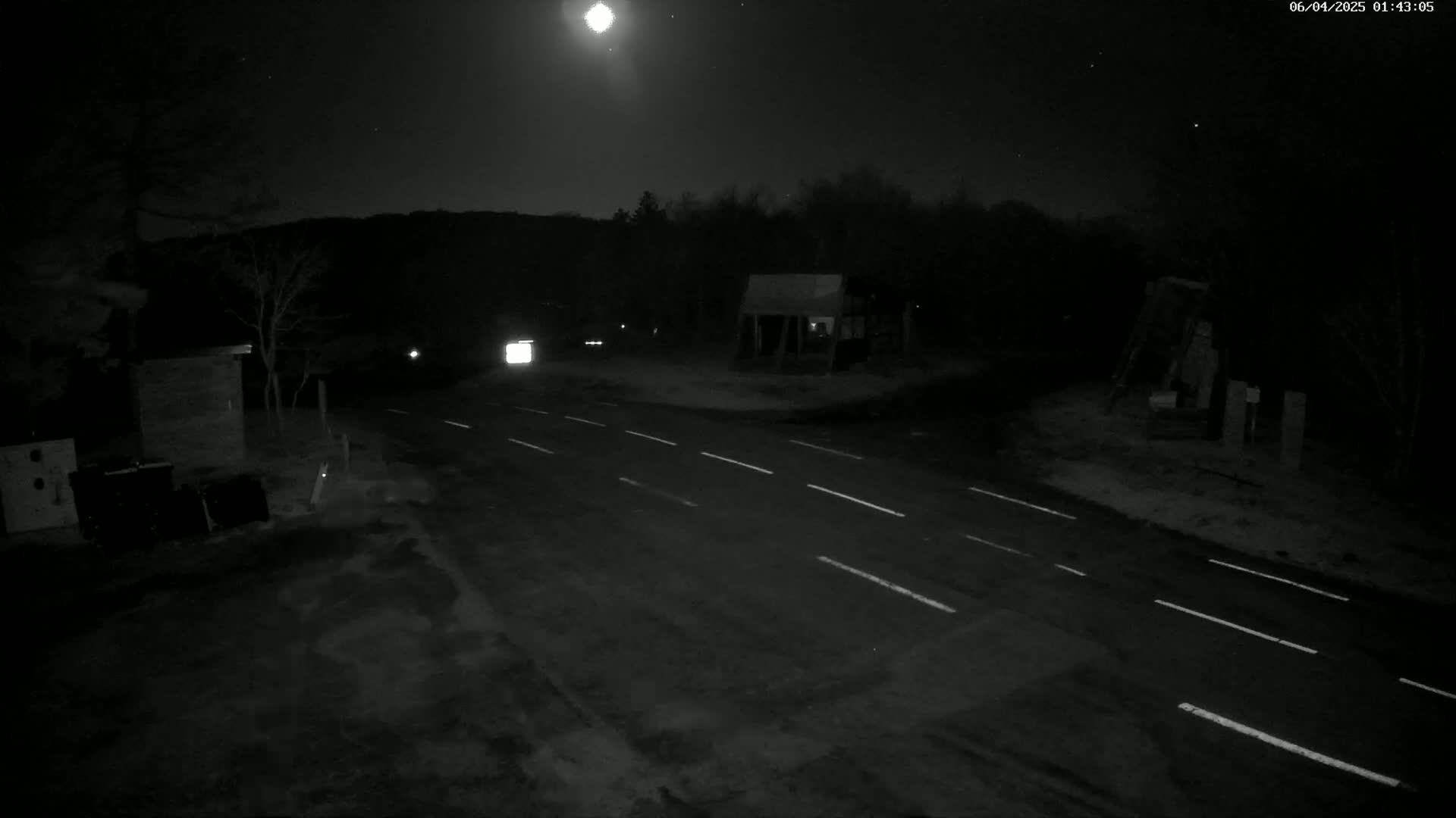 A nighttime, moonlit view of a rural road intersection with sparse vegetation and small structures.