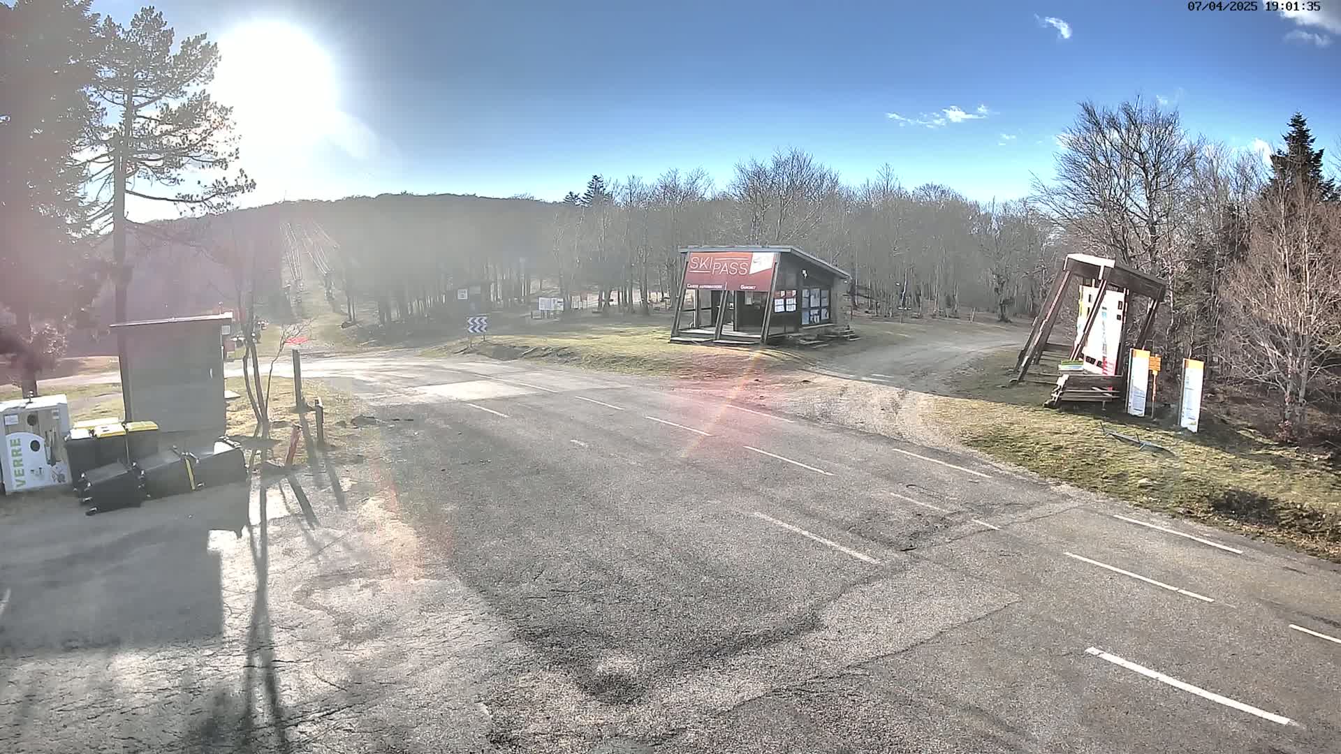 A sunny day at a ski resort parking lot with a small ski pass building and several informational signs visible, surrounded by bare trees.