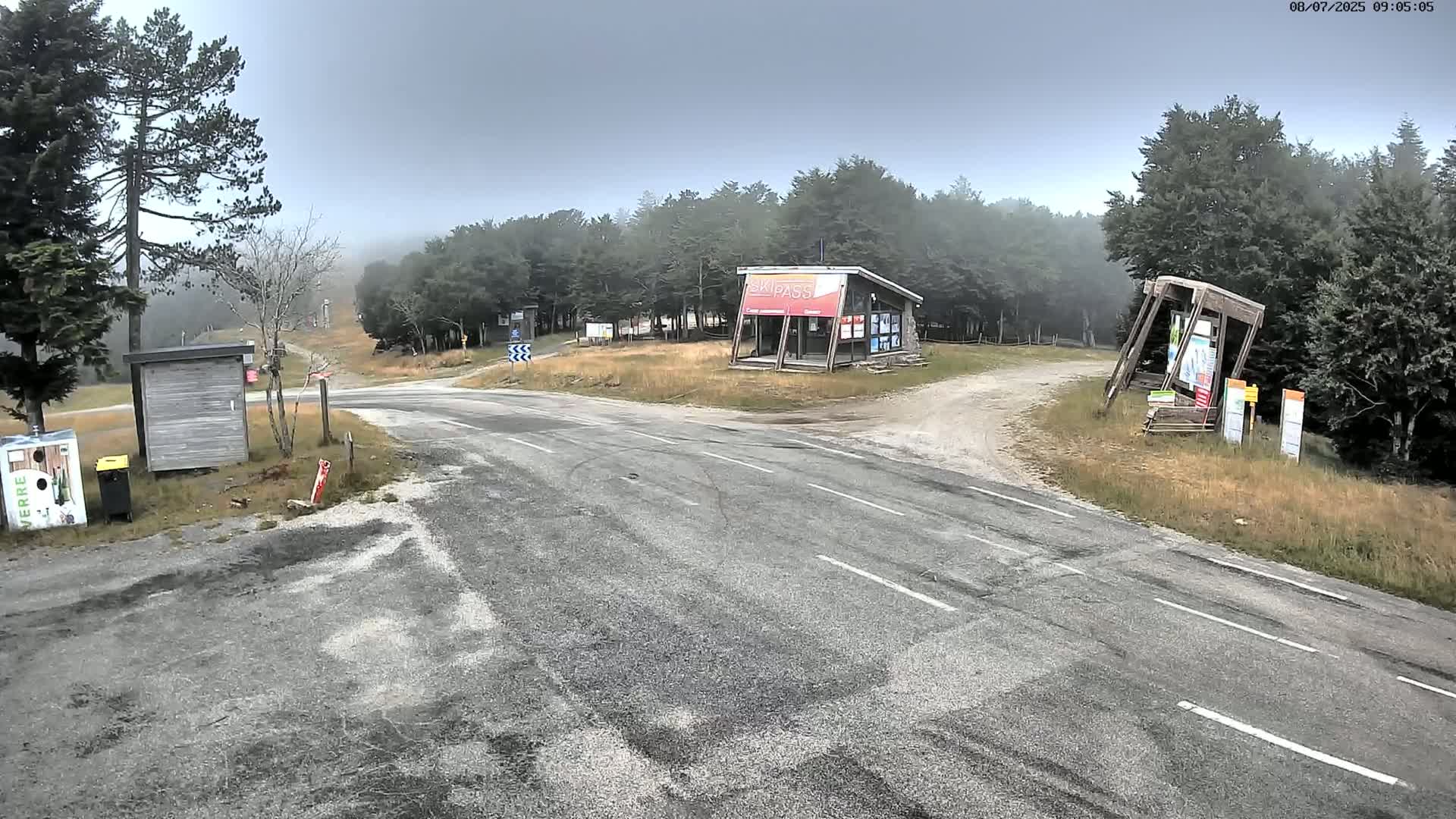 A foggy mountain road intersection with a small building and several informational kiosks is surrounded by trees.