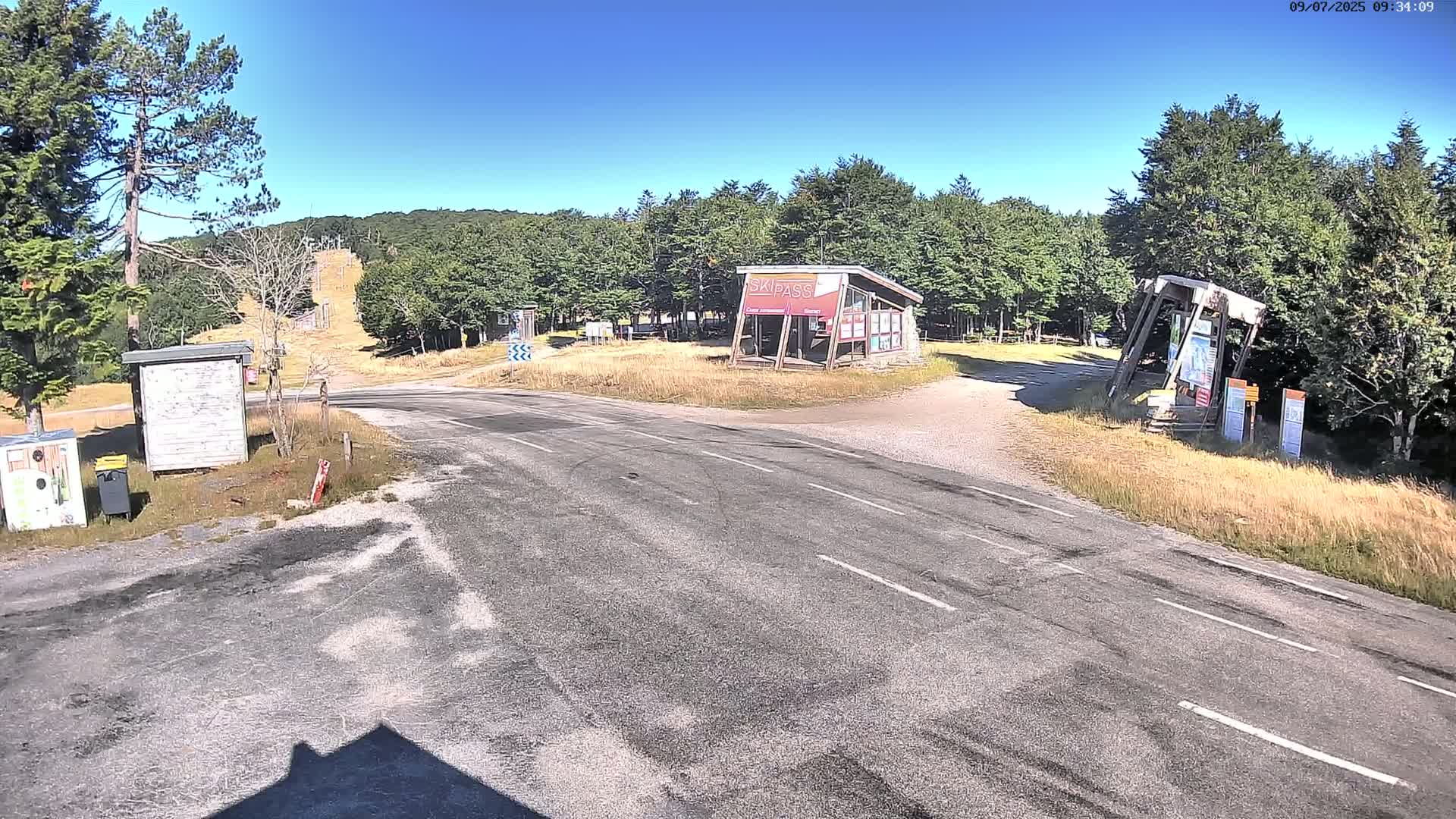 A paved road forks at an intersection near a ski lift in a sunny, wooded area.
