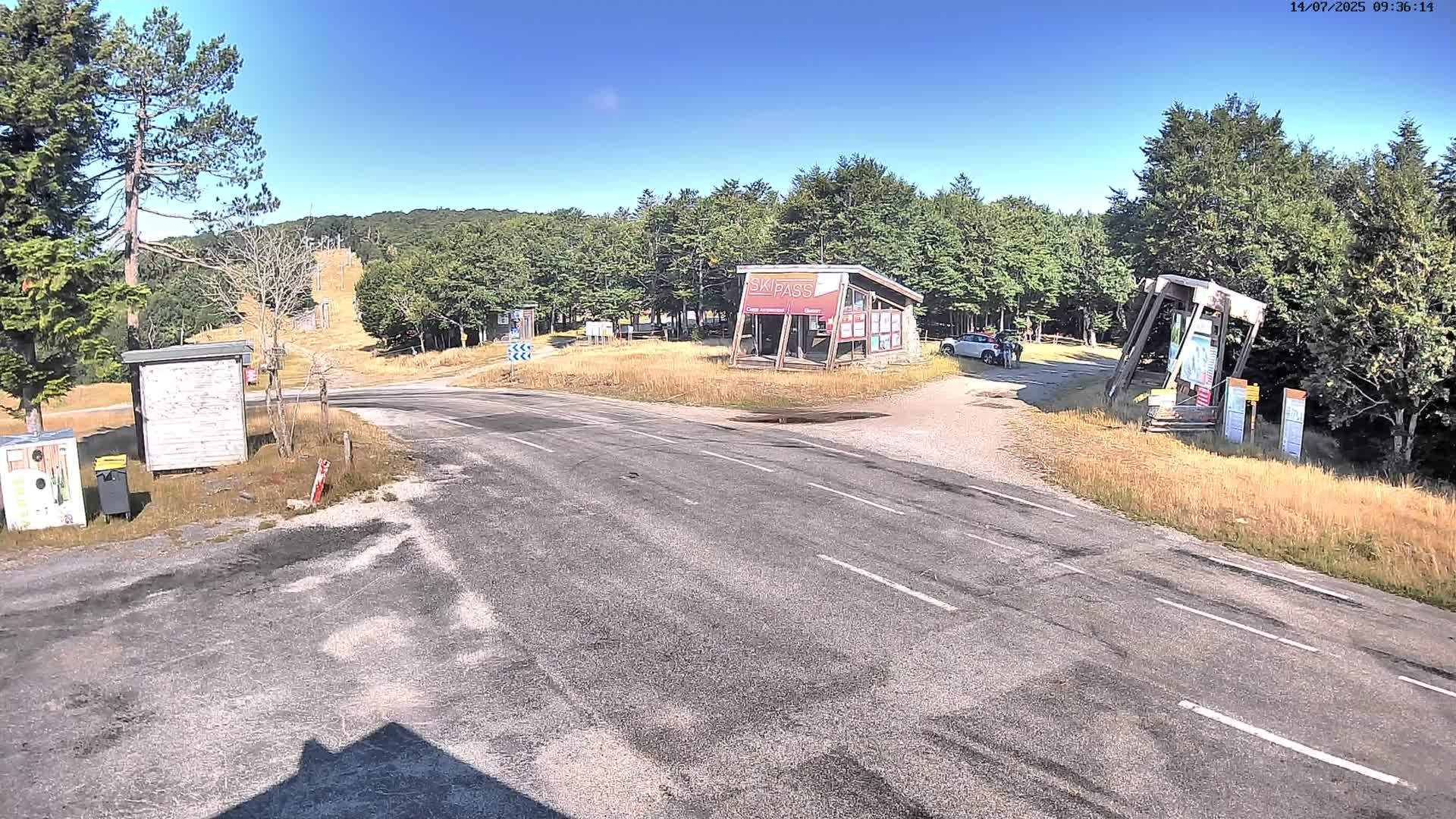 A paved road forks at a mountaintop intersection near a ski lift and several small buildings, under a clear blue sky.