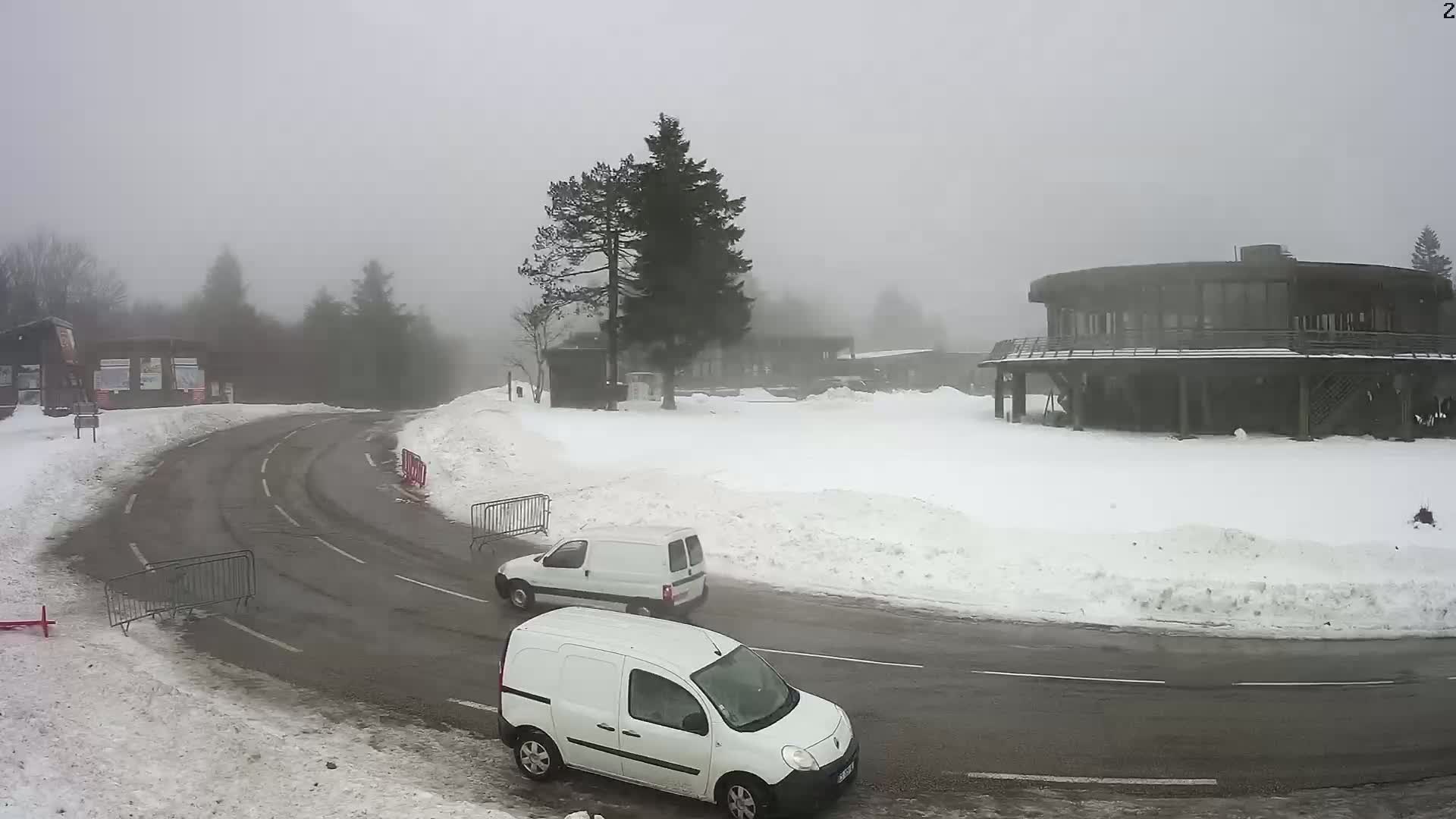 Two white vans are parked on a snow-covered road curving around a large building in foggy conditions.