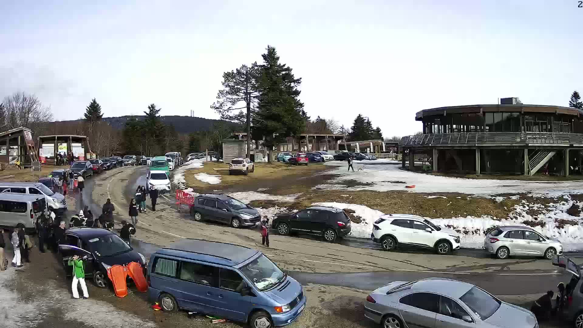 A parking lot and roadway filled with cars and people in a snowy, partially sunny area near a large building.