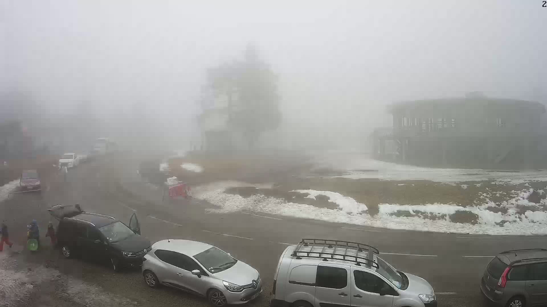 A foggy parking lot with several cars and a few people, some patches of snow and ice are visible near the roadside.