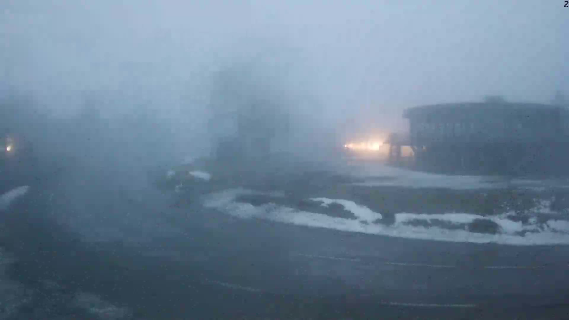 A foggy, snowy scene shows a road curving around a building with patches of snow and a few vehicles barely visible in the heavy weather.