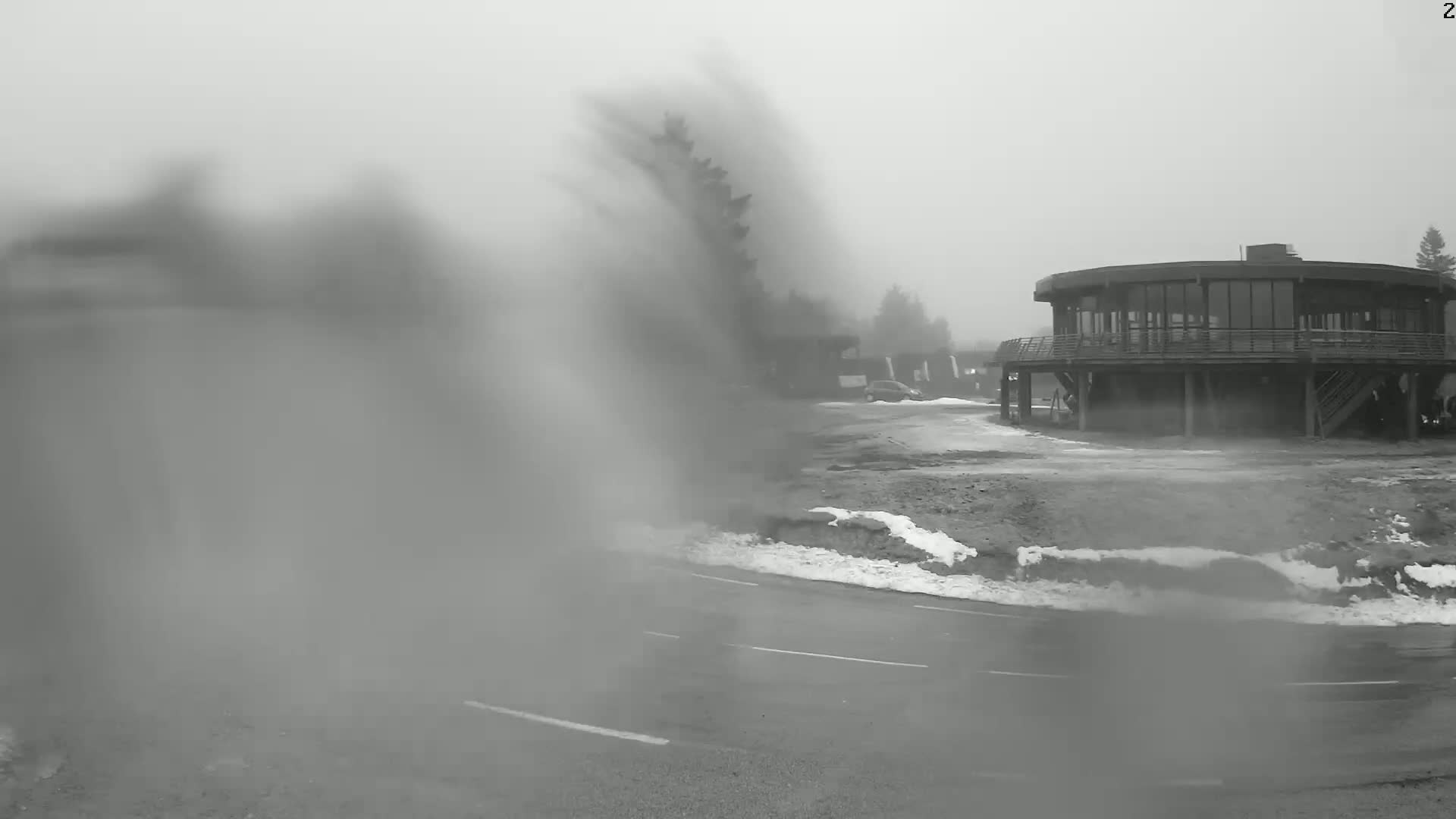 A blurry, foggy view shows a circular building with a deck and stairs, partially obscured by blowing snow, near a road with patches of snow.