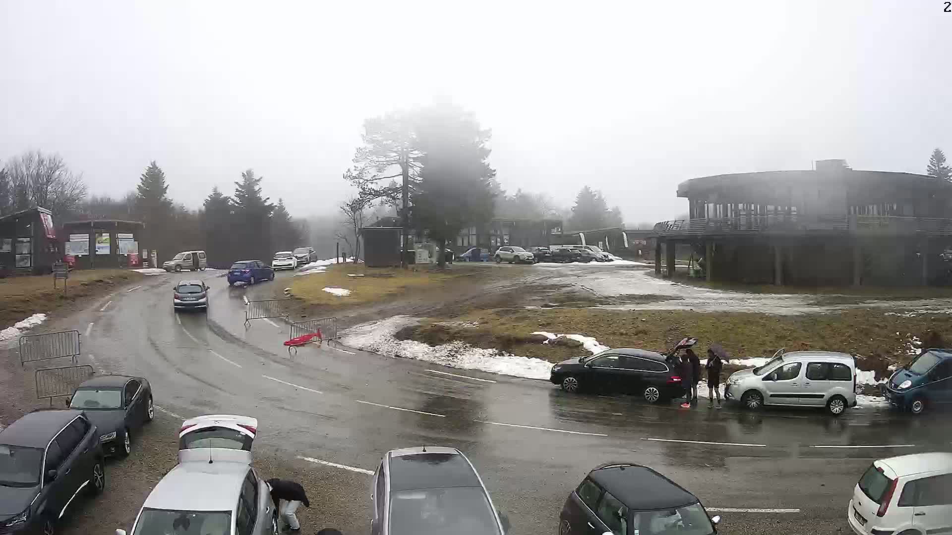 A wet, foggy parking lot and roadway near a building surrounded by trees, with several cars parked and driving.