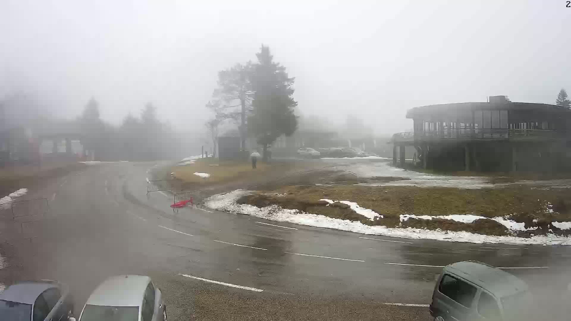 A foggy, wet roadway curves around a building and a small parking lot with patchy snow and several parked cars.