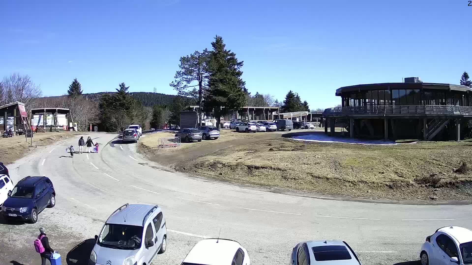 A sunny day reveals a parking lot with several cars near a circular building and a family walking with children.