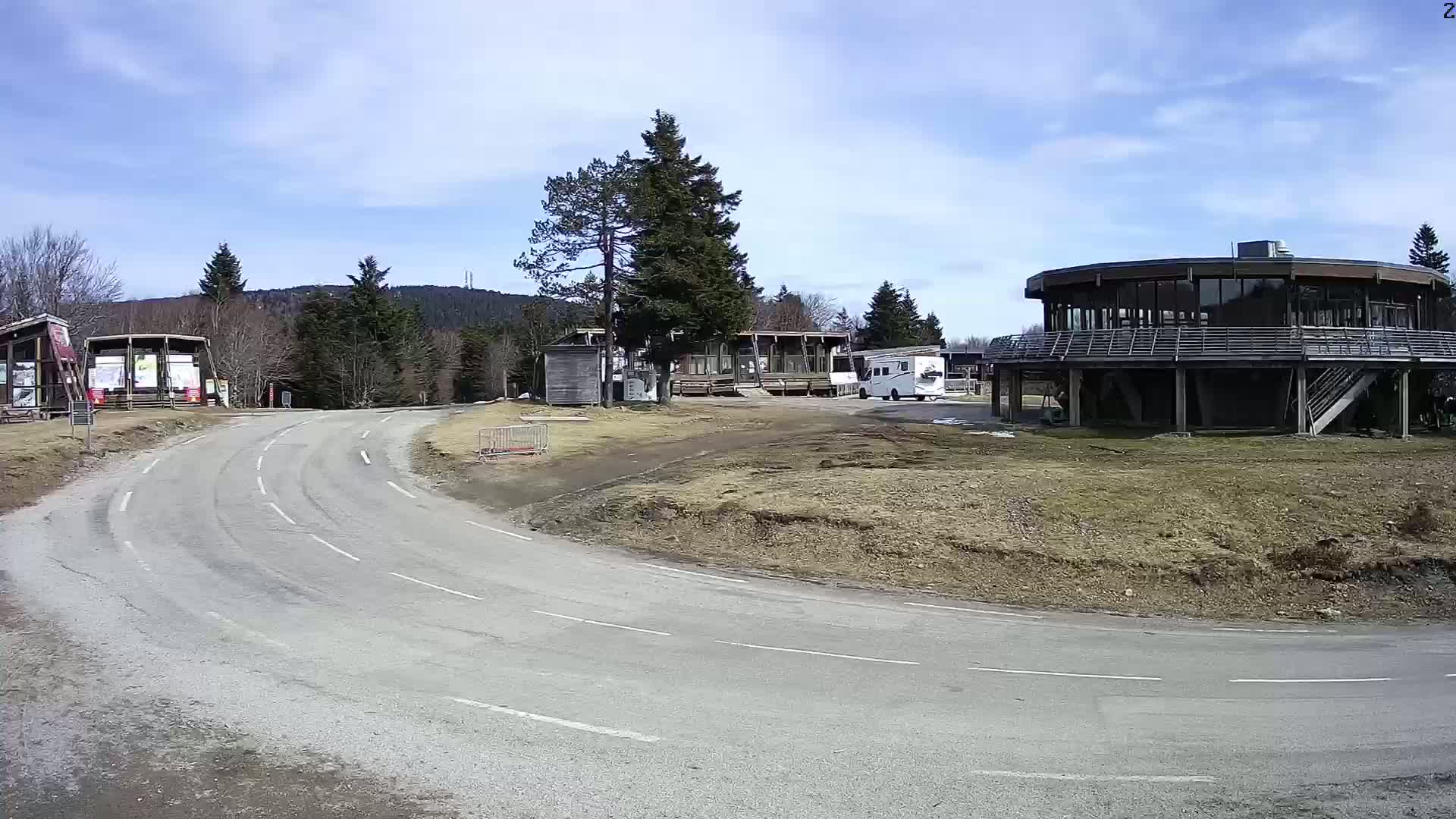 A curving road leads to several buildings, including a large circular one, in a mountainous area under a mostly sunny sky.