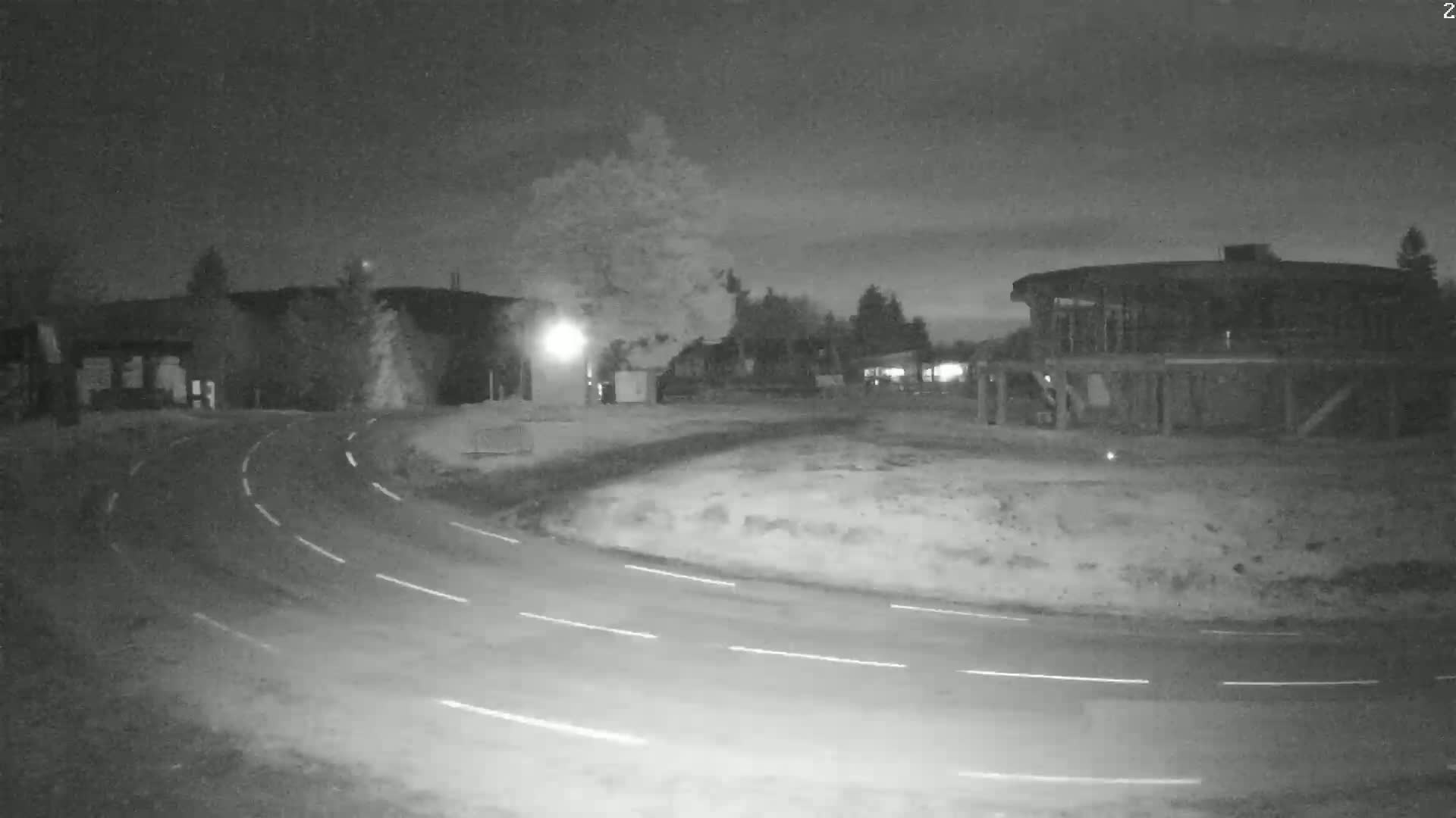 A nighttime, low-light image shows a curved road with white lane markers winding past buildings and trees under a dark, possibly cloudy sky.