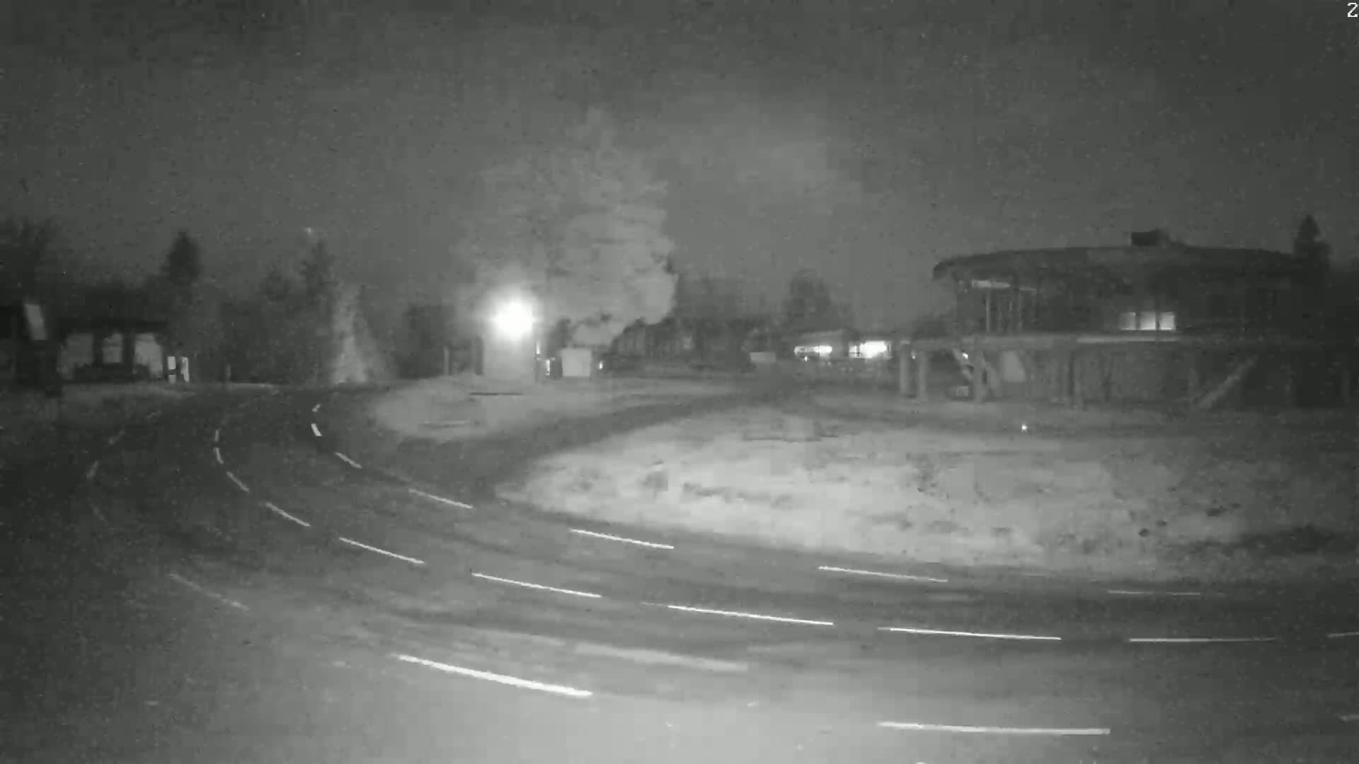 A nighttime, snow-covered scene shows a curved roadway with faint tire tracks leading to a building and several trees under a dark sky.