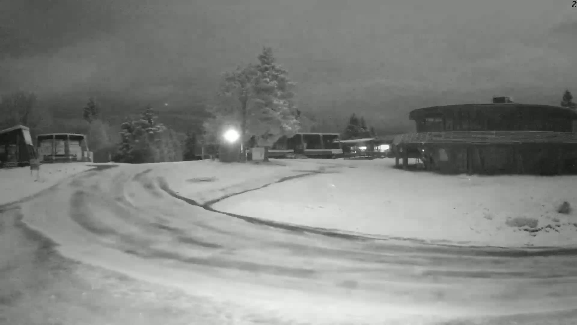 A snow-covered roadway curves around a building and several vehicles under a dark, overcast sky.