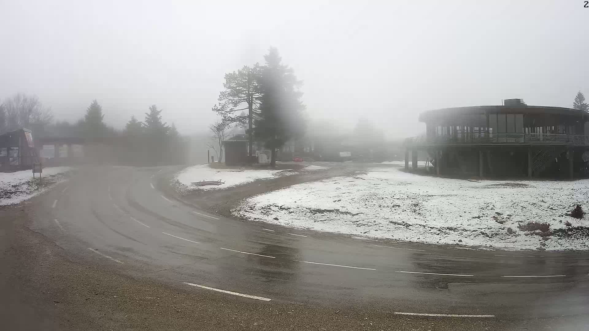 A curving road with tire tracks winds through a foggy, snowy landscape, past a circular building and other structures partially obscured by the heavy mist.