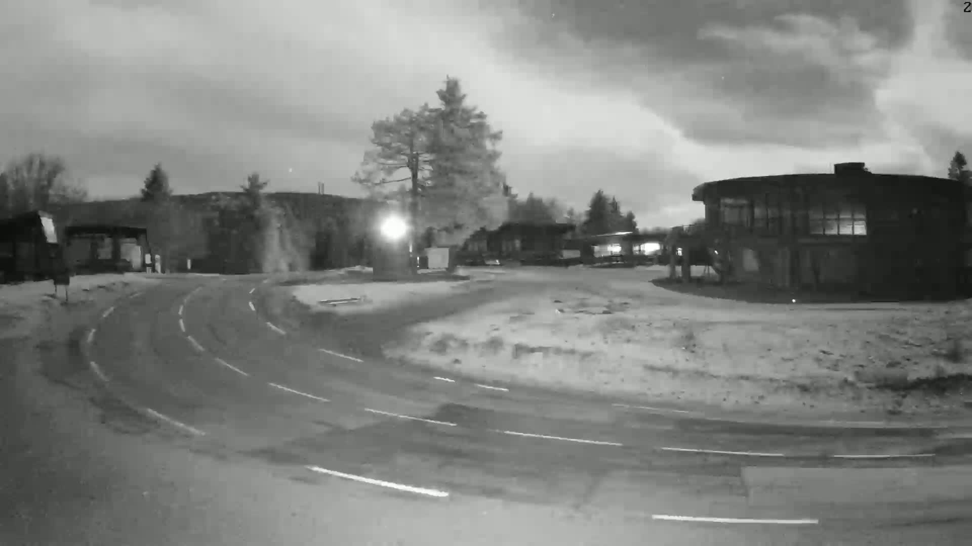 A nighttime, snow-covered scene shows a curved road leading to several buildings under a cloudy sky.