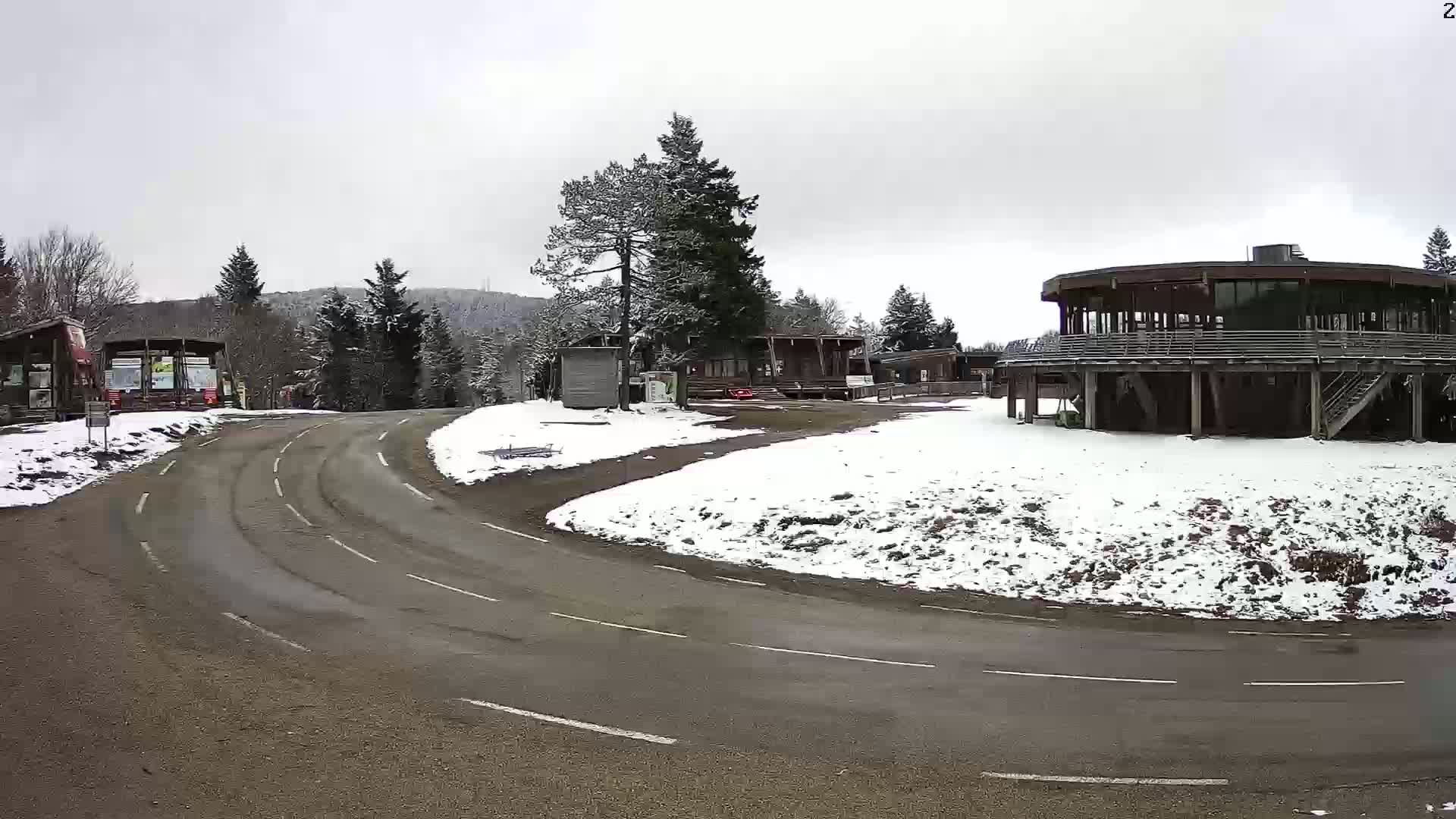A curved road leads to a snow-covered area with several buildings, including a large circular structure, surrounded by pine trees under an overcast sky.