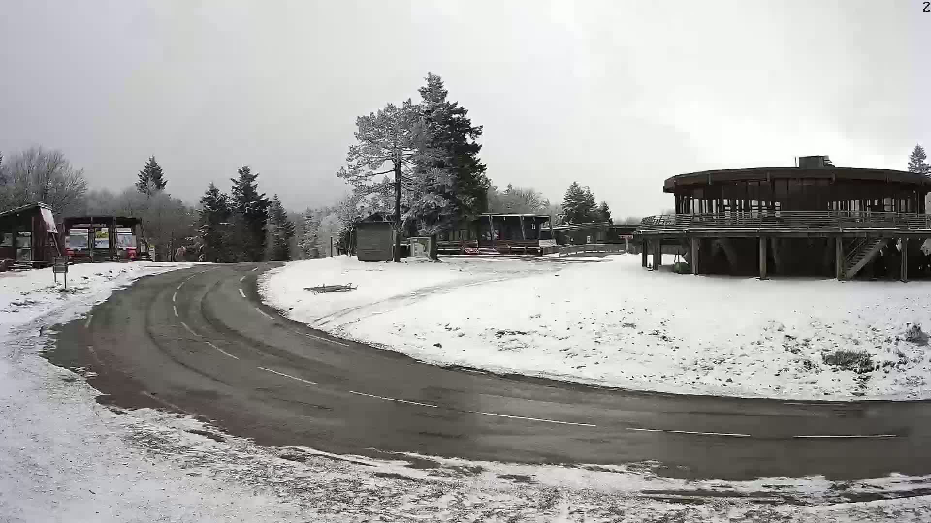 A snow-covered roadway curves around a cluster of buildings nestled in a snowy, wooded area under an overcast sky.