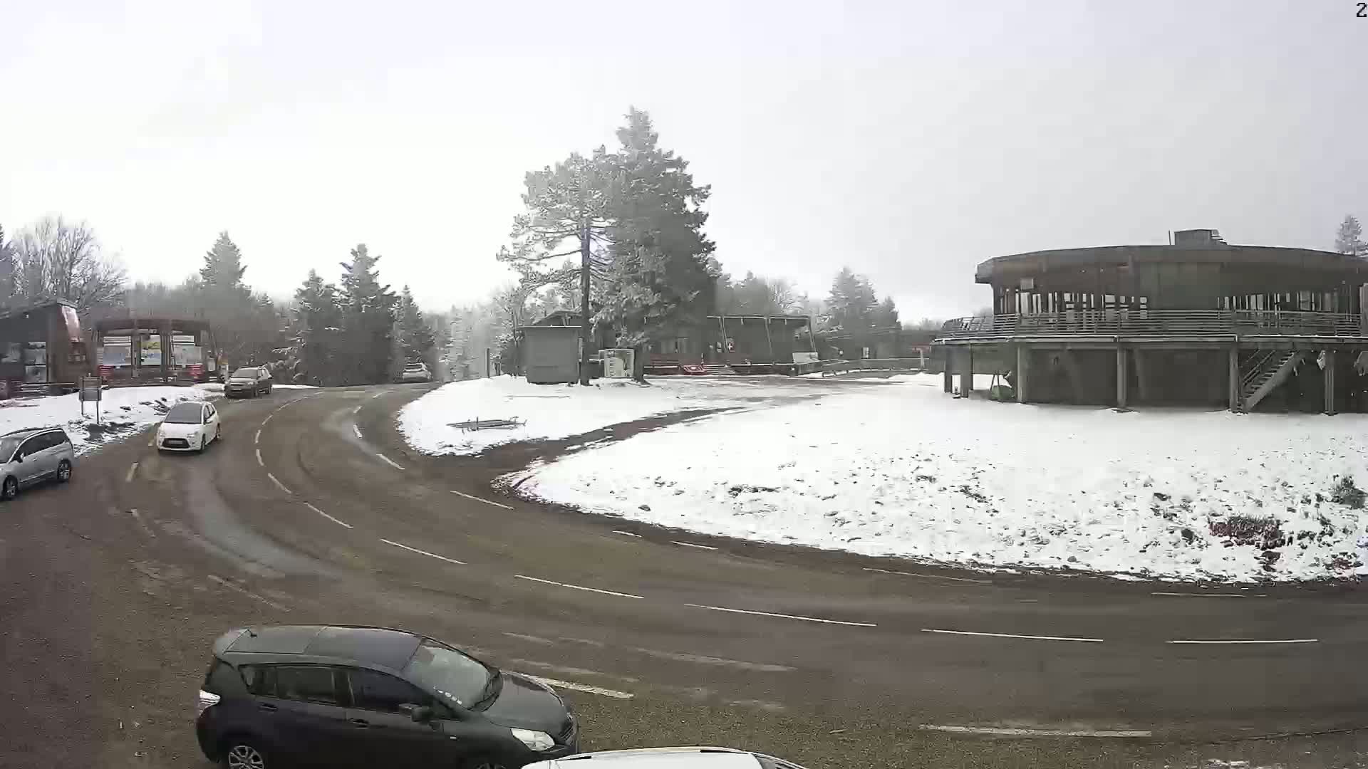 A snow-covered roadway curves around a circular building and a small parking area, with several cars visible in the scene under a light snowfall.
