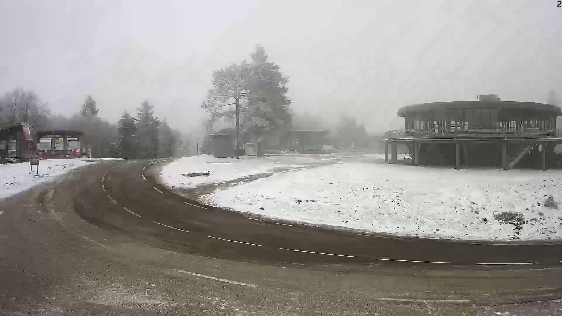 A snow-covered roadway curves around a circular building and several smaller structures in foggy, snowy weather.