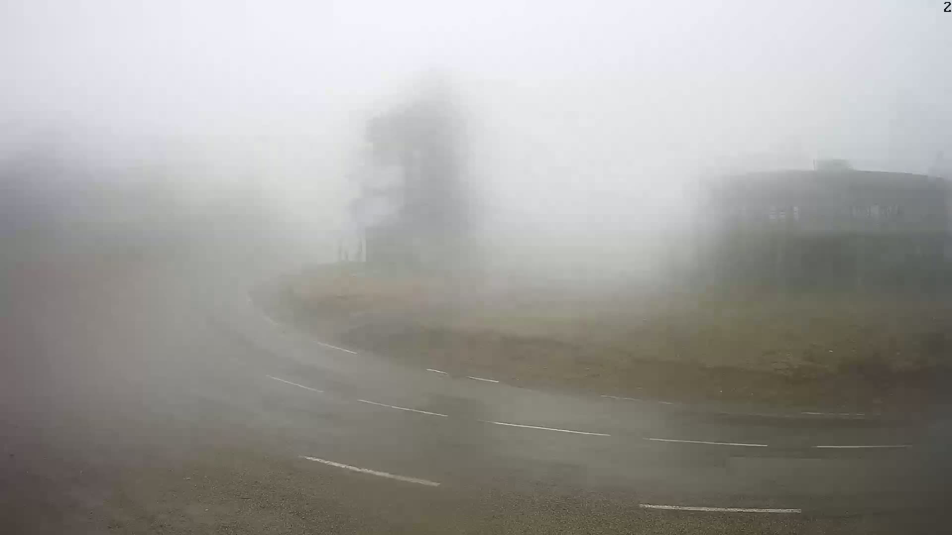 A wet, foggy road curves past a building and a tall structure, partially obscured by heavy fog.