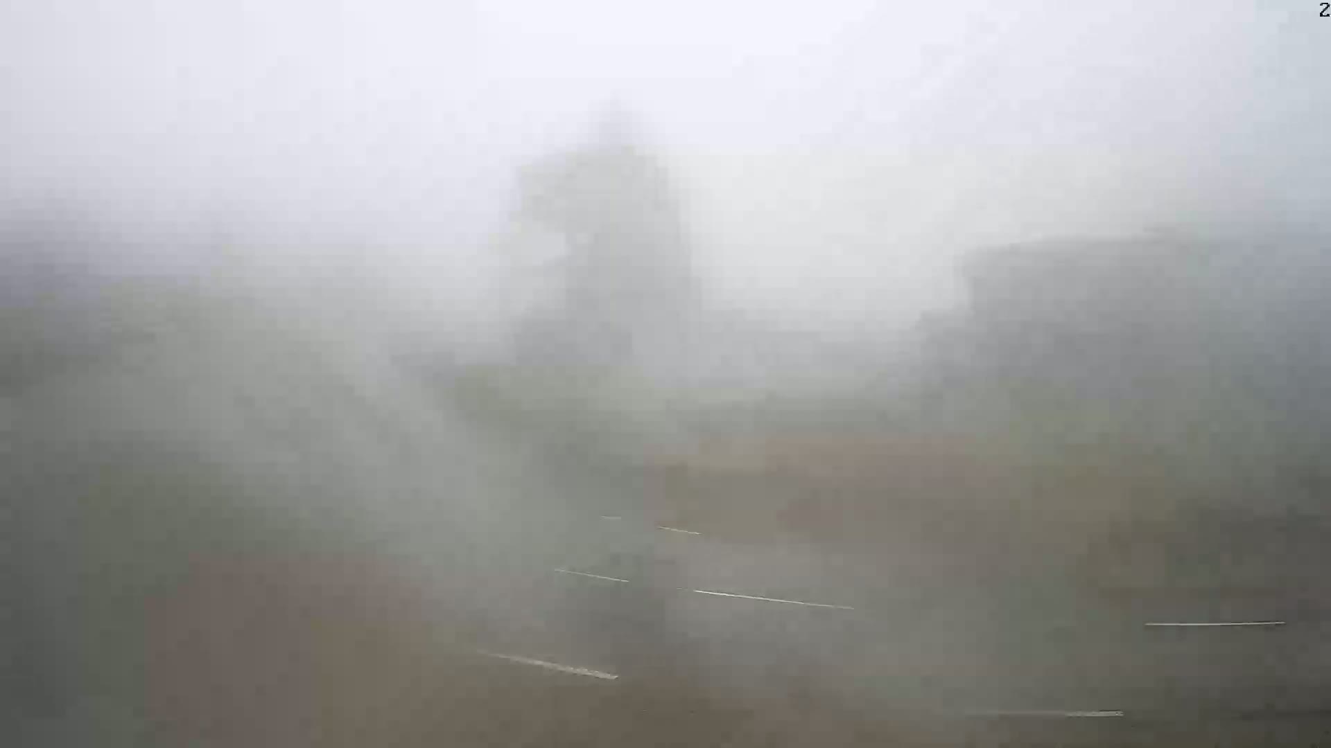A foggy outdoor scene shows indistinct shapes of buildings and vegetation, with a road visible in the foreground.