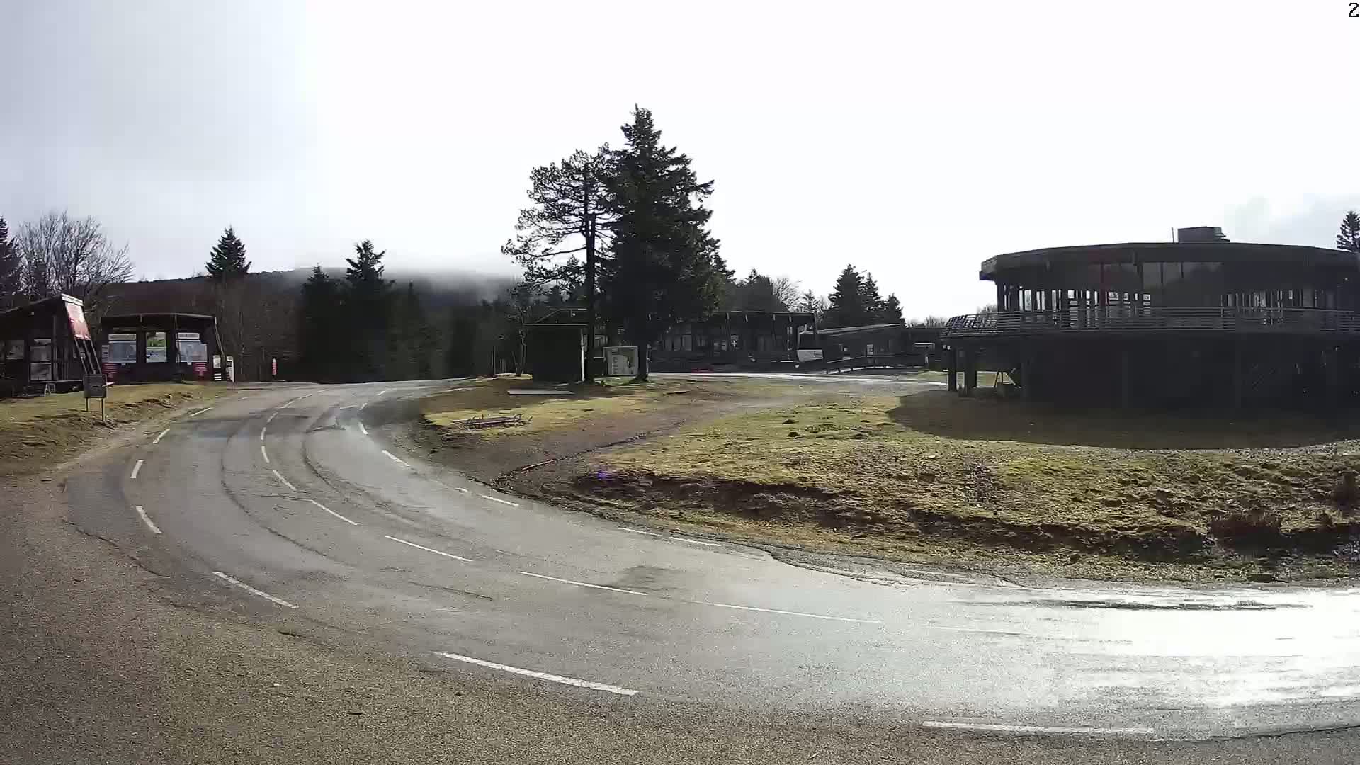 A wet asphalt road curves around a grassy area towards a circular building and several smaller structures nestled among evergreen trees under a bright, hazy sky.