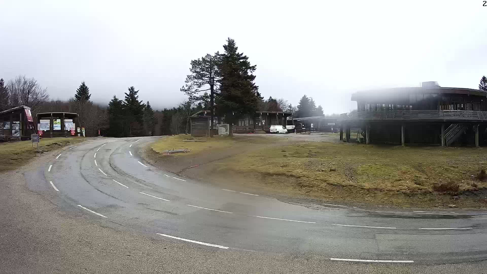 A wet, curving road leads to a cluster of low-lying buildings in a wooded area under an overcast sky.