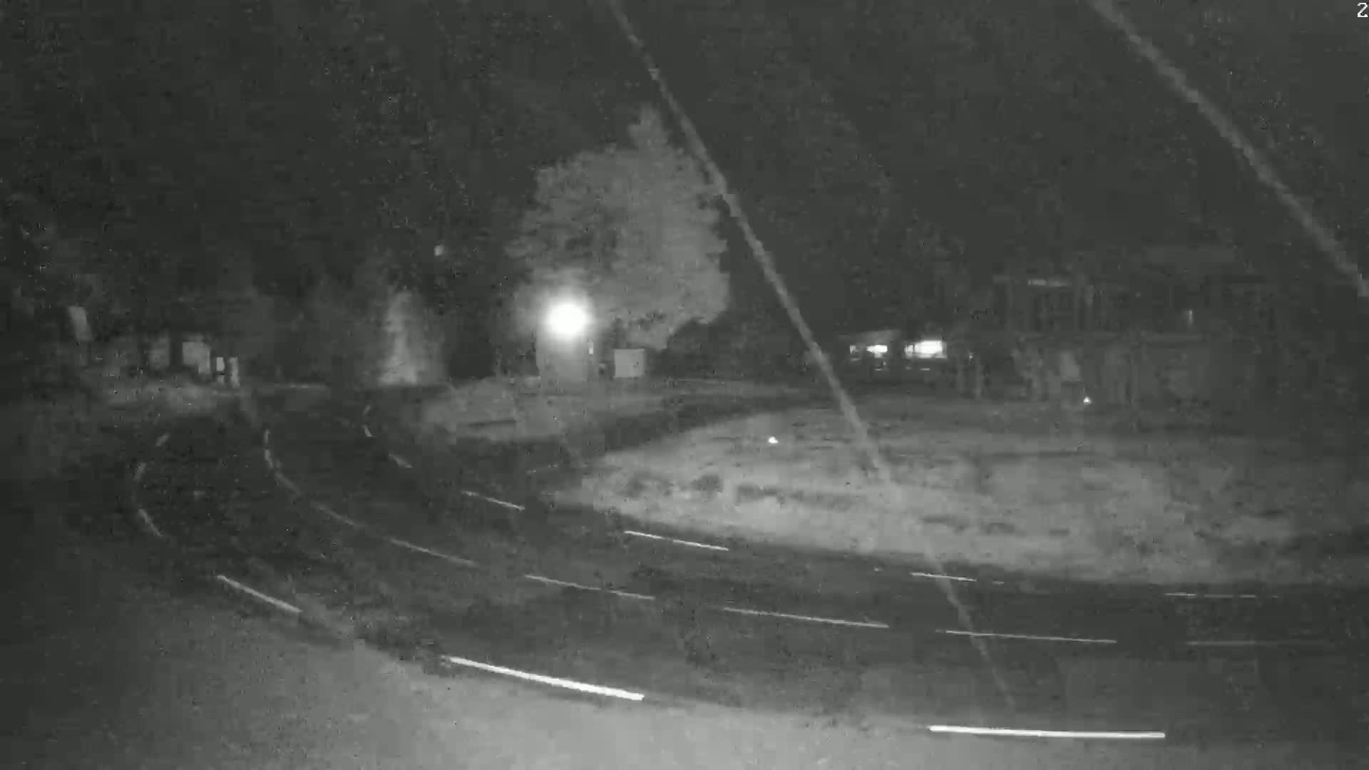 A nighttime, low-light view of a roundabout with minimal traffic, surrounded by trees and buildings.