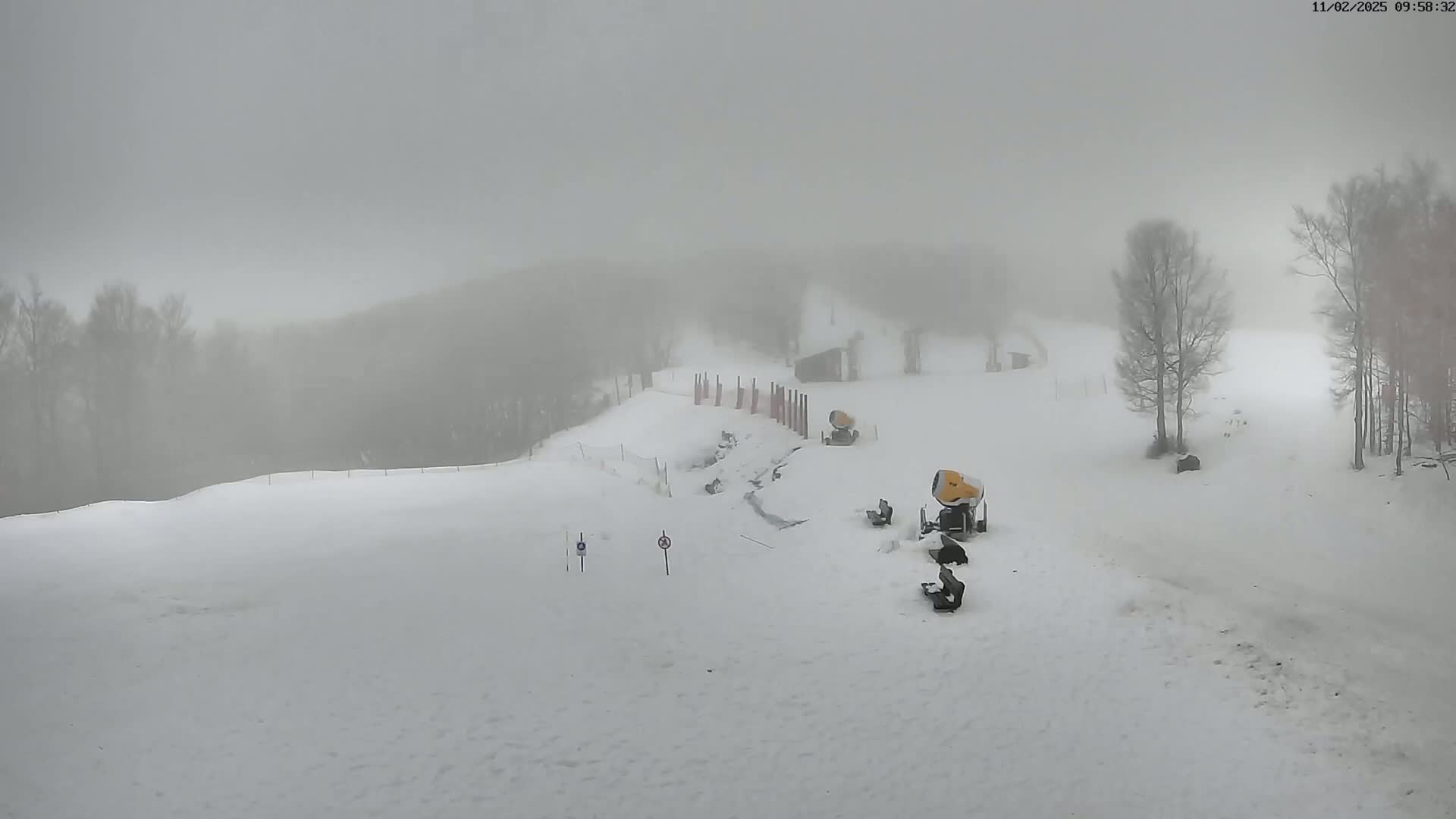 A snowy ski slope is shrouded in fog, with snowmaking machines and some minimal equipment visible near the bottom of the hill.