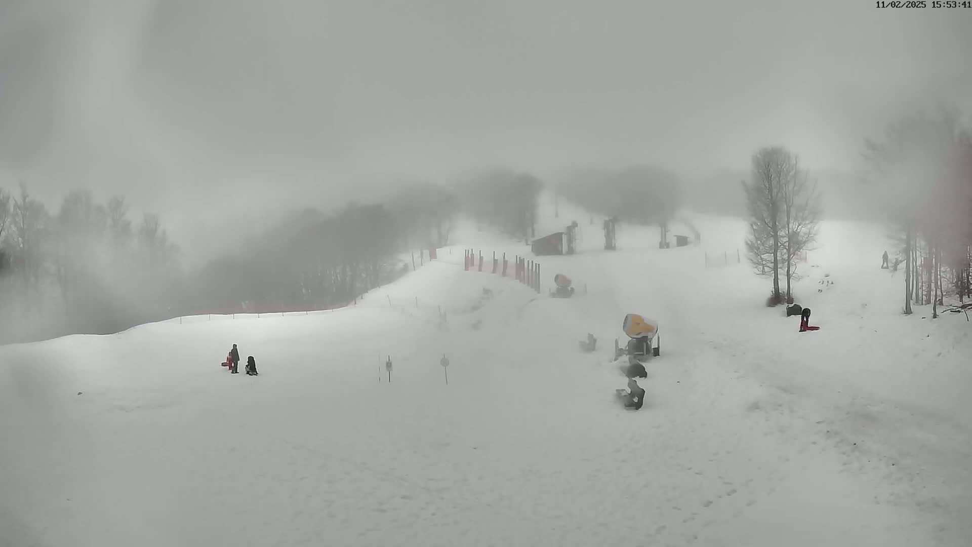 A snowy ski slope is shrouded in dense fog, with a few people and snow-grooming machines visible.